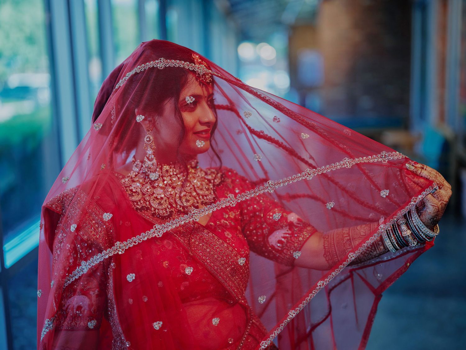 Indian bride in red lehenga and dupatta with gold jewelry poses elegantly at her wedding ceremony.