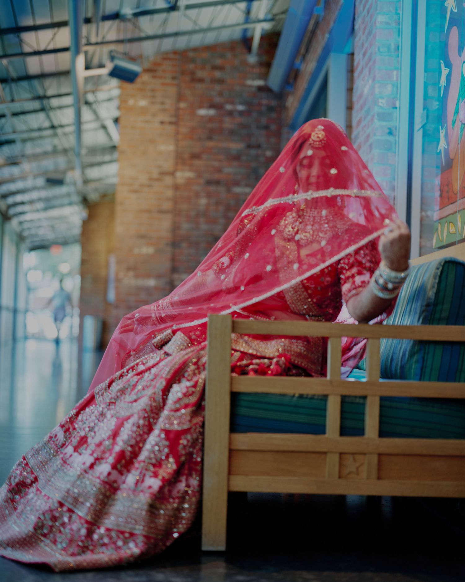 Indian bride in red bridal lehenga seated on wooden bench with embroidered dupatta draped over face.