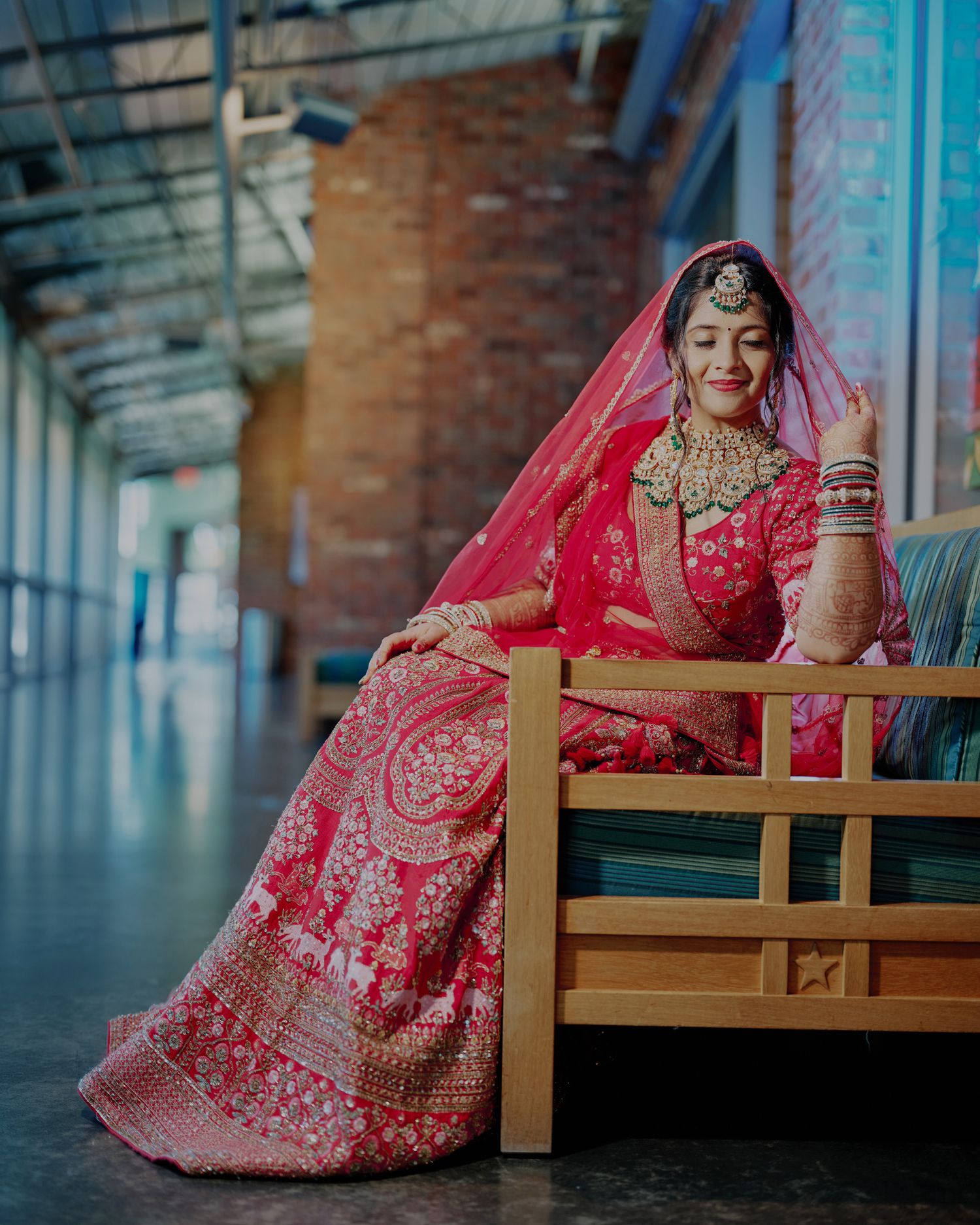 Bride in red bridal lehenga with intricate embroidery and dupatta seated on wooden bench at Indian wedding.