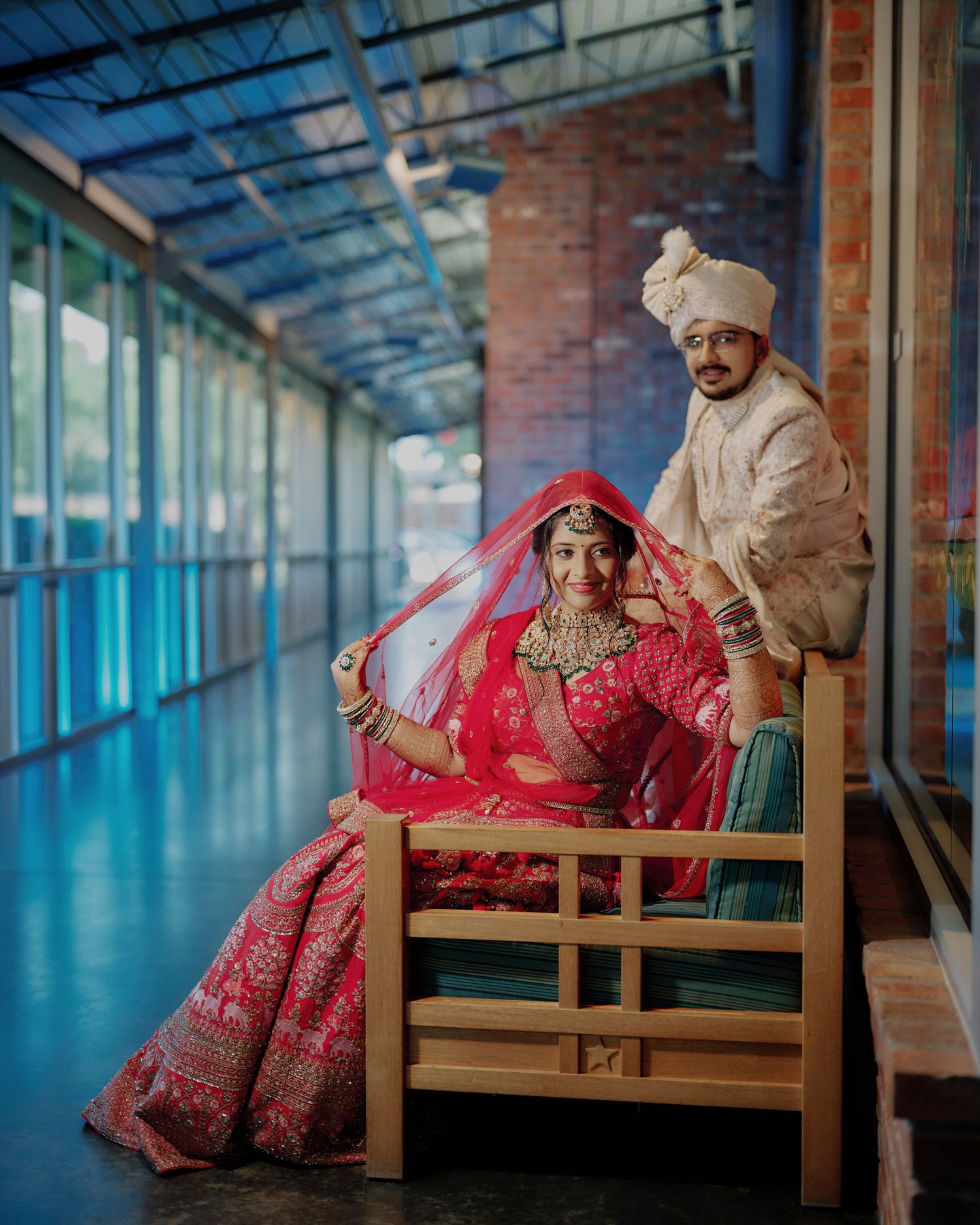Indian bride and groom in traditional red and ivory wedding attire posing on wooden bench in corridor.