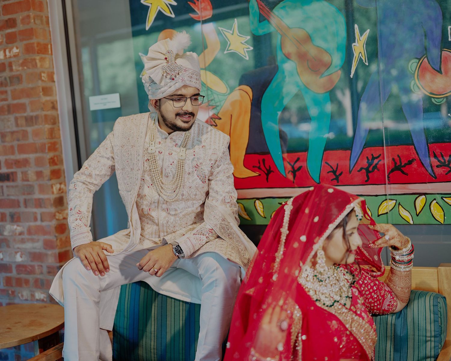 Indian groom in white sherwani and turban sits beside bride in red bridal lehenga during traditional wedding ceremony.