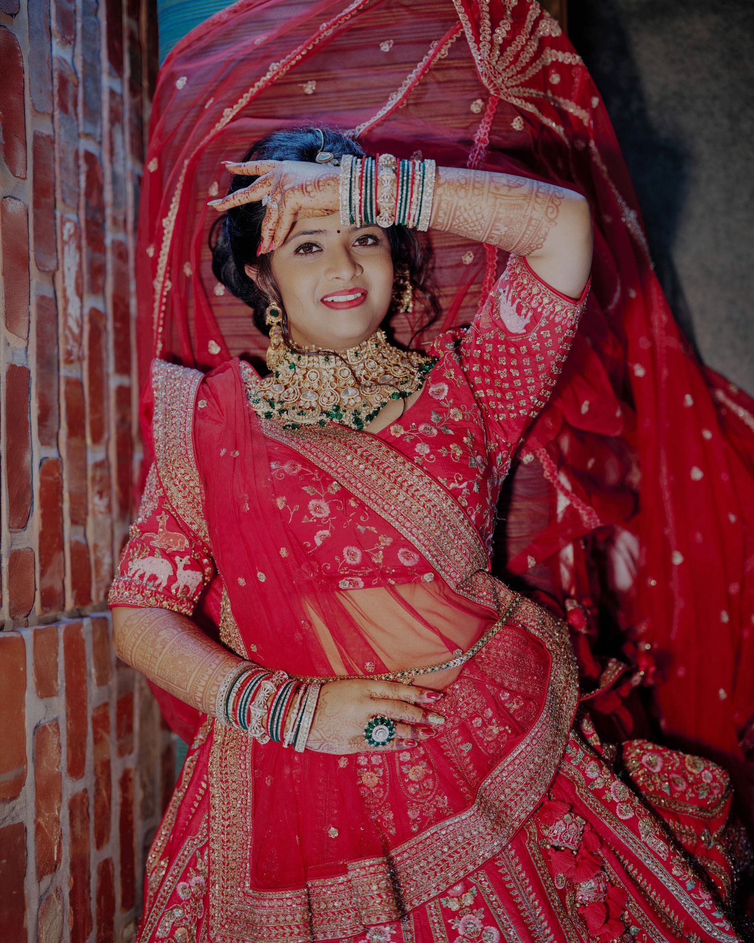 Indian bride in red lehenga with gold jewelry, mehndi hands, and red dupatta posing gracefully.