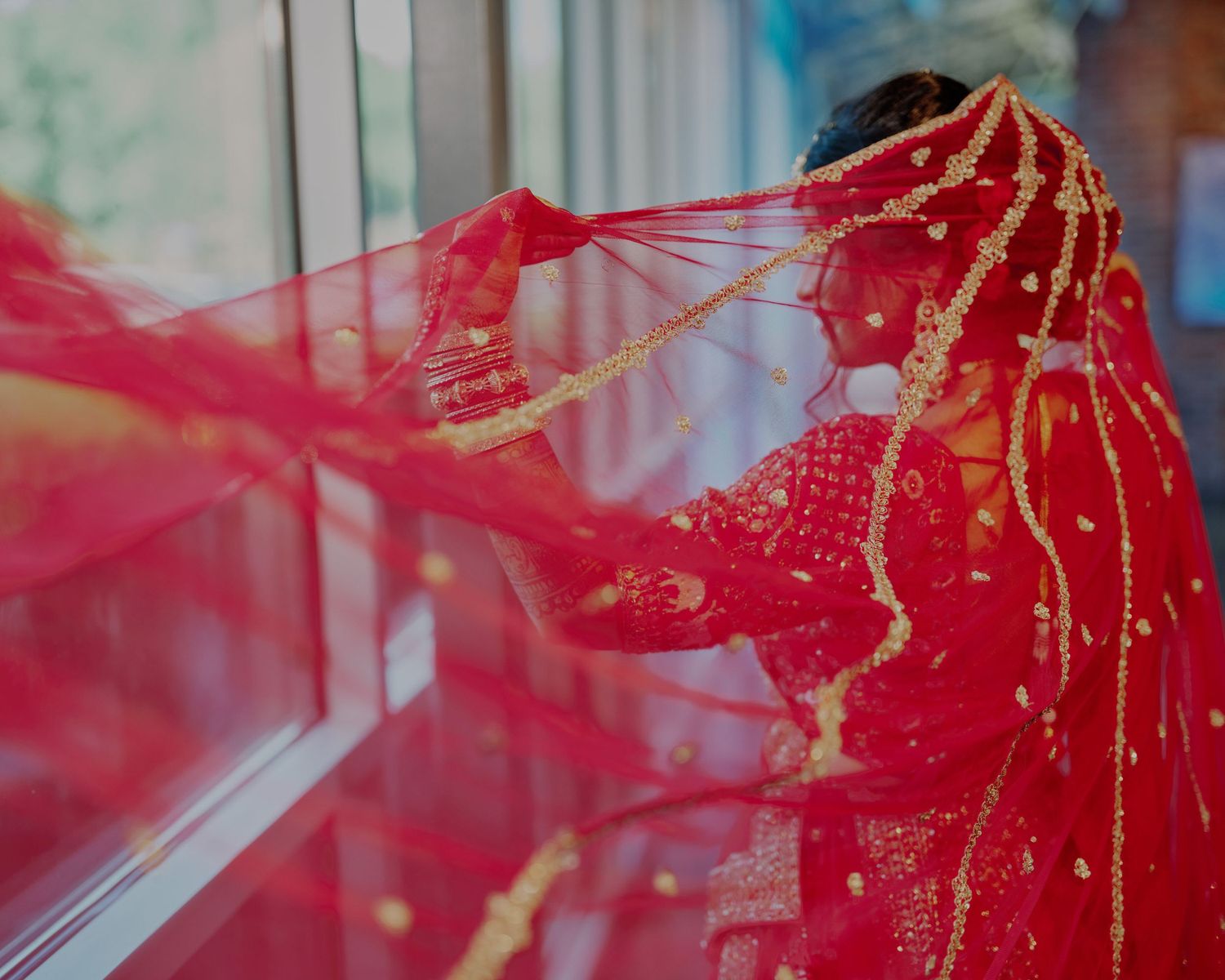A bride in a vibrant red bridal outfit with gold embroidery, her dupatta flowing gracefully near a window.