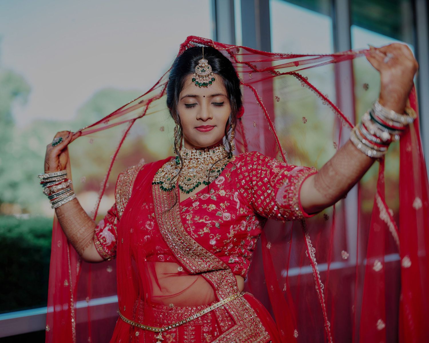 A bride in a red lehenga and gold jewelry holds her dupatta veil gracefully during a traditional Indian wedding.