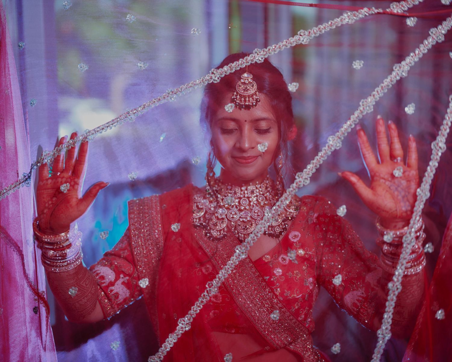 Indian bride in red lehenga with henna hands and bridal jewelry posing behind decorative crystal curtain.