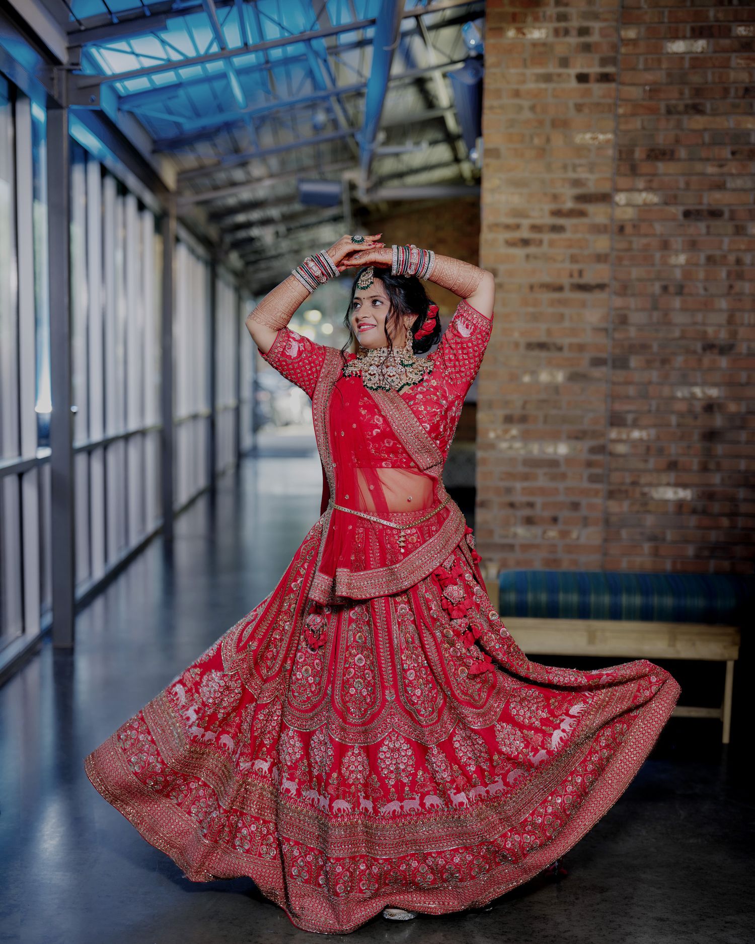 Bride twirling in a stunning red embroidered lehenga with traditional jewelry at an indoor venue.