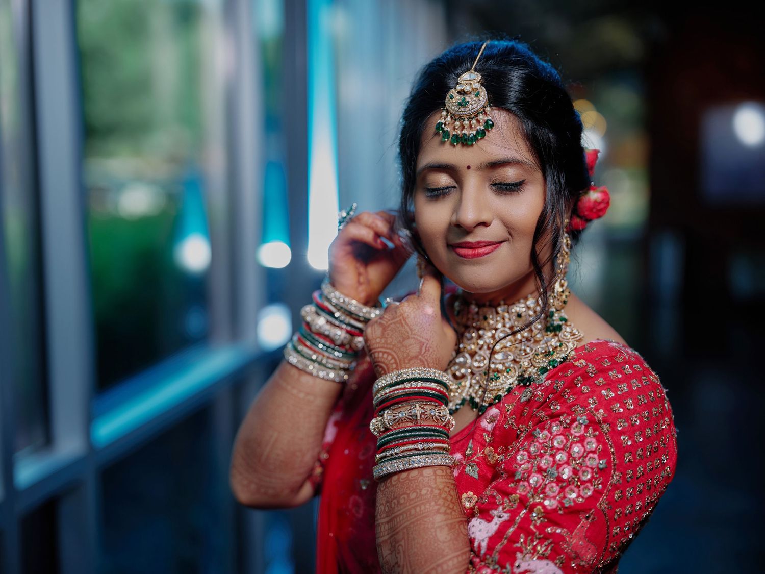 Indian bride in red lehenga adjusting earring wearing traditional gold jewelry and bangles at wedding.