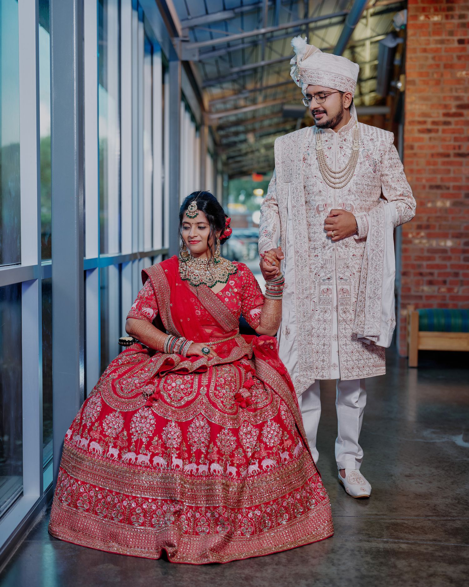 Indian bride in red lehenga sits as groom in white sherwani holds her hand at wedding ceremony.