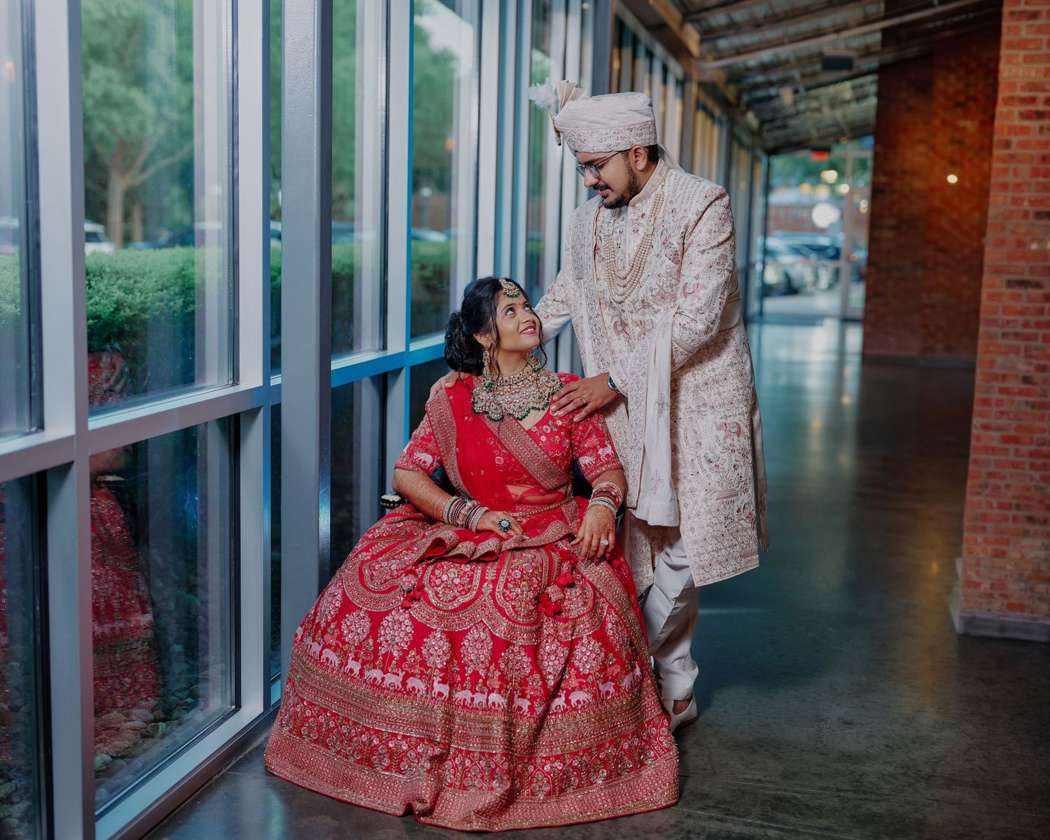 Indian couple in traditional wedding attire sharing romantic moment in glass corridor wedding portrait.