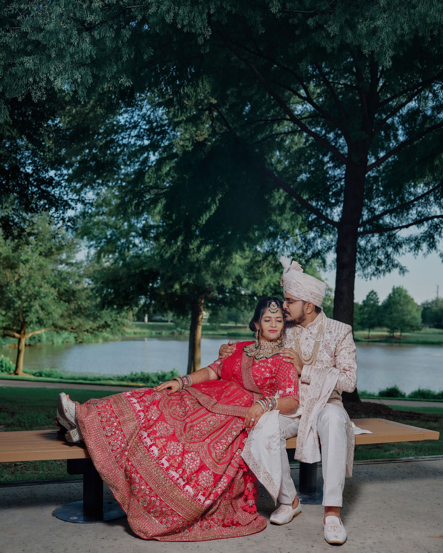 Indian bride in red lehenga and groom in sherwani sit together by a lake in lush park setting.