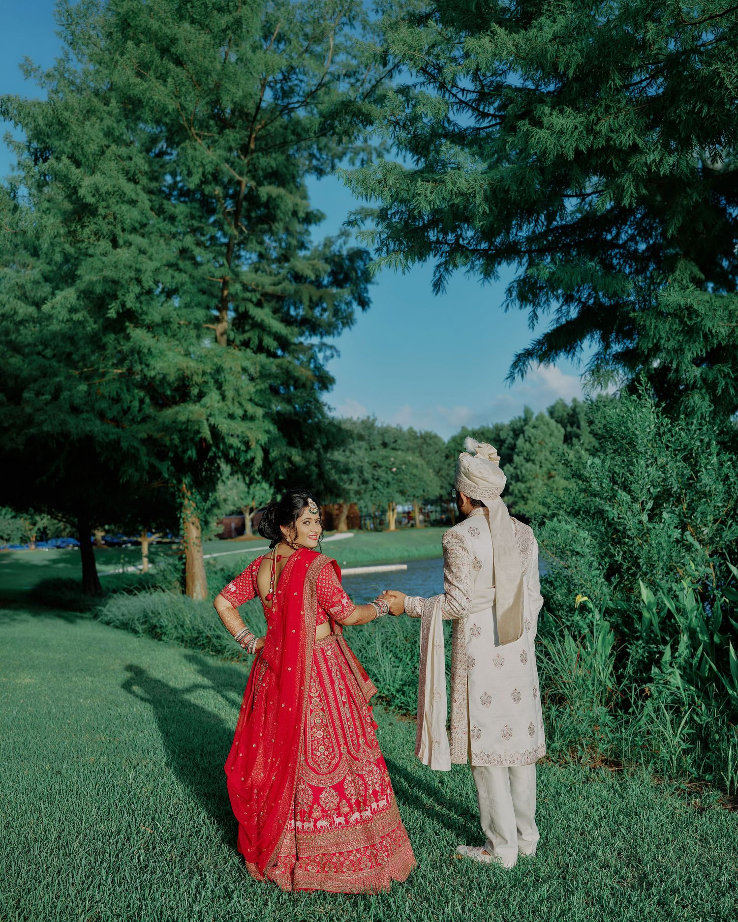 Indian couple in traditional wedding attire holding hands in a lush green garden, bride in red lehenga, groom in cream sherwani.