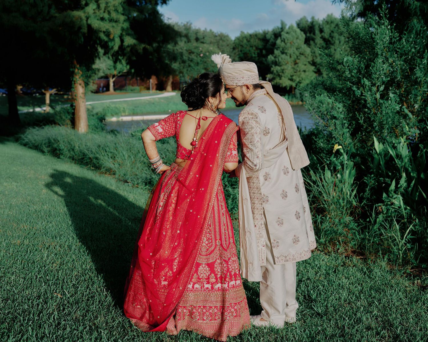 Indian bride in red lehenga and groom in beige sherwani share intimate moment in lush garden wedding photo.