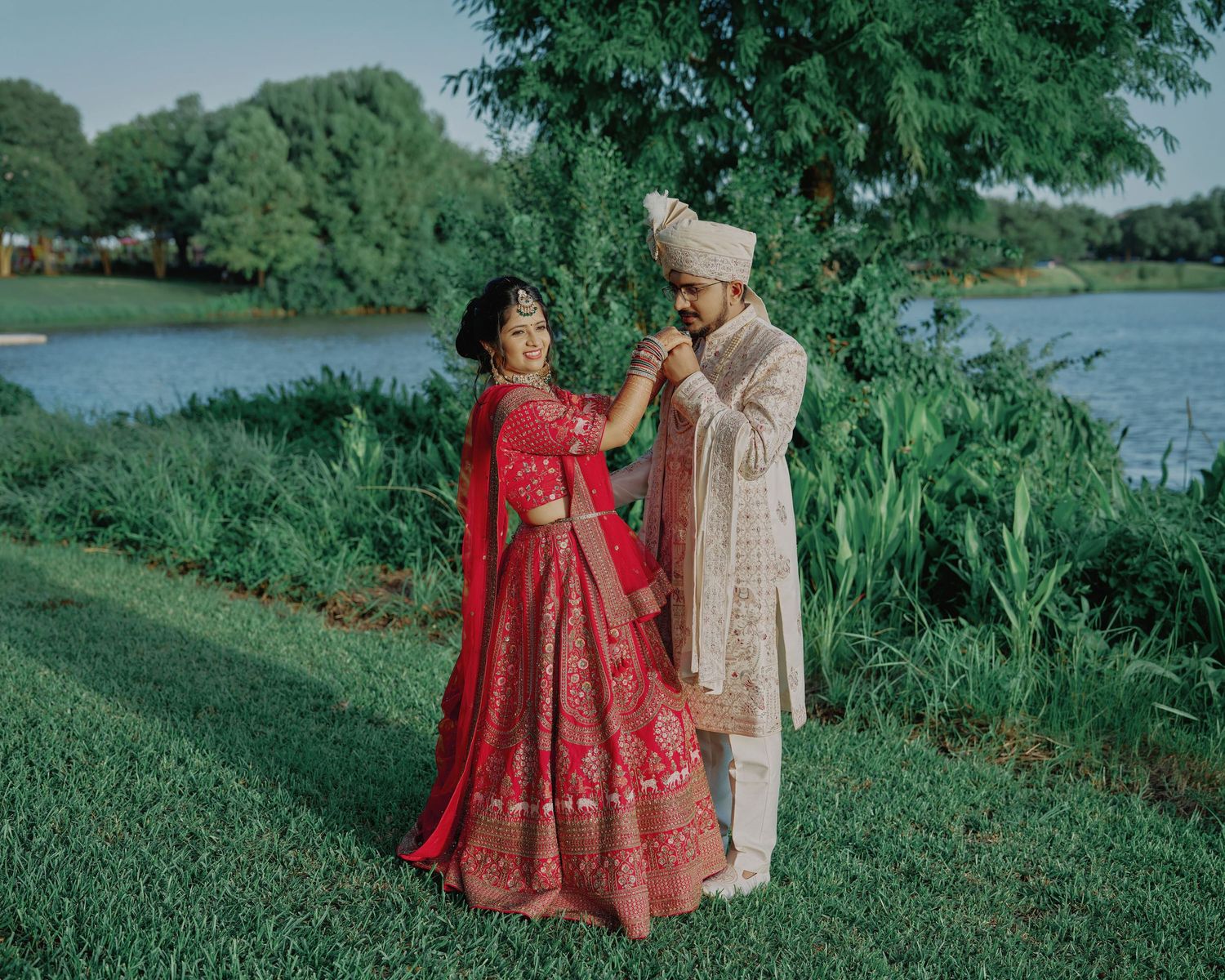 Indian couple in traditional wedding attire share a romantic moment by a serene lakeside garden.