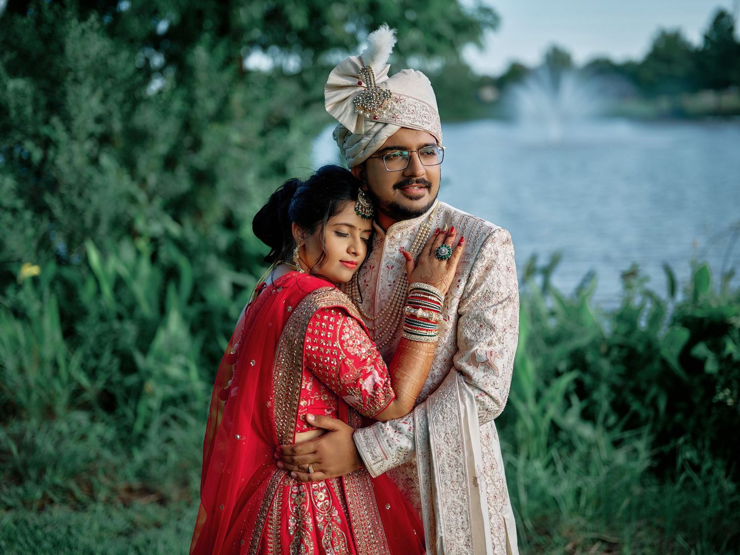 Indian couple in traditional wedding attire embracing near a lake, bride in red lehenga, groom in ivory sherwani.