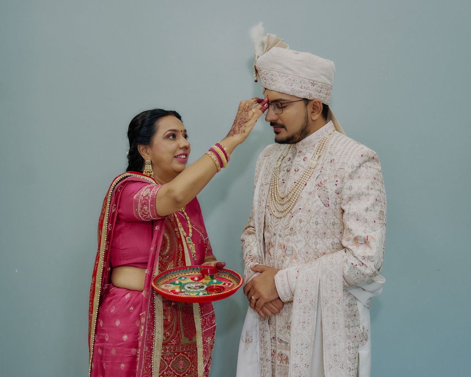 Indian woman in pink saree applying tilak to groom in white sherwani during wedding ritual.