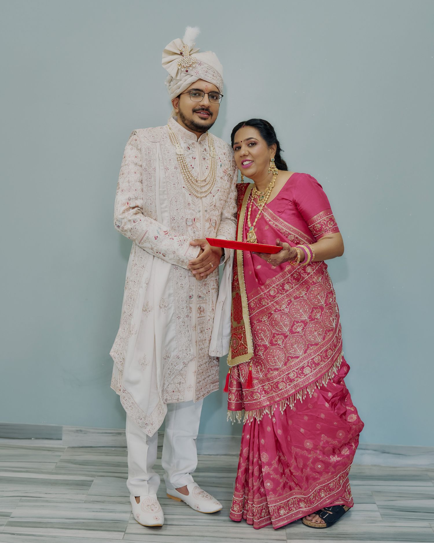 Indian bride in pink silk saree and groom in white sherwani holding red puja thali together.