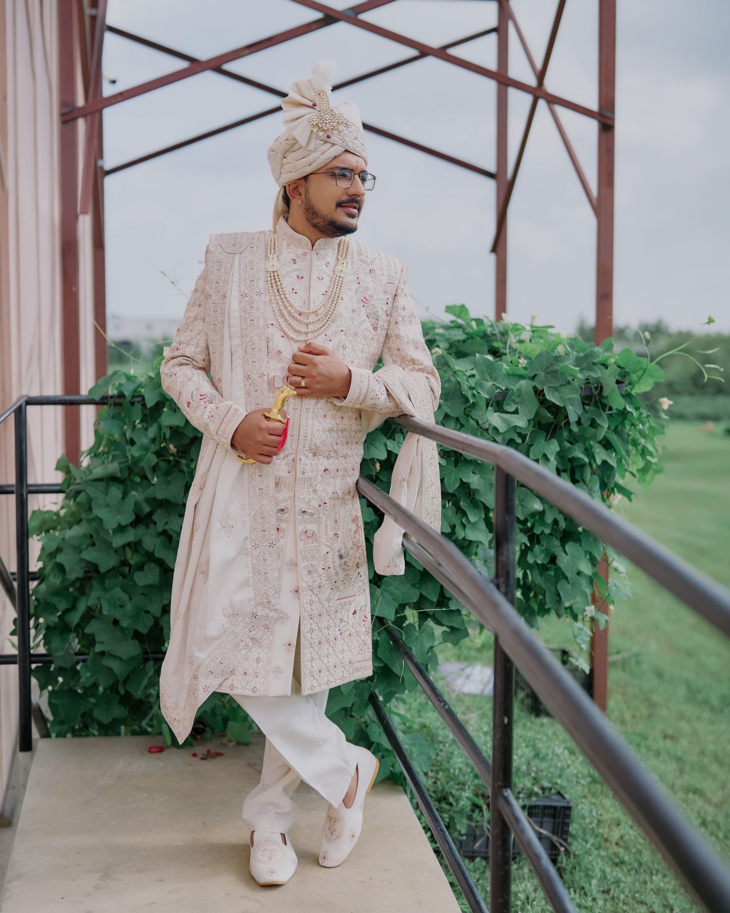 Indian groom in cream embroidered sherwani and turban standing on balcony surrounded by greenery.