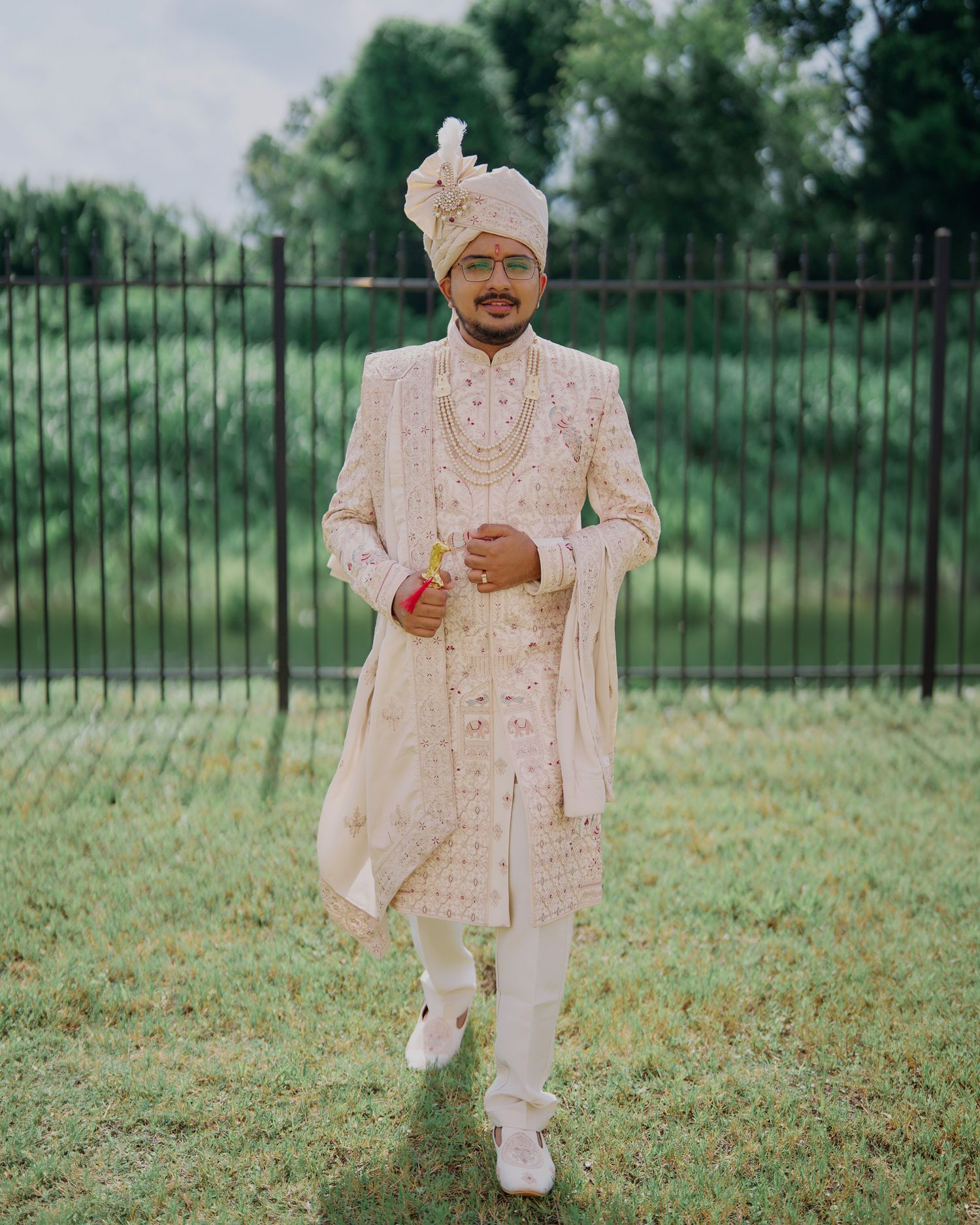 Man in traditional Indian sherwani and turban stands on grass near iron fence for wedding ceremony.