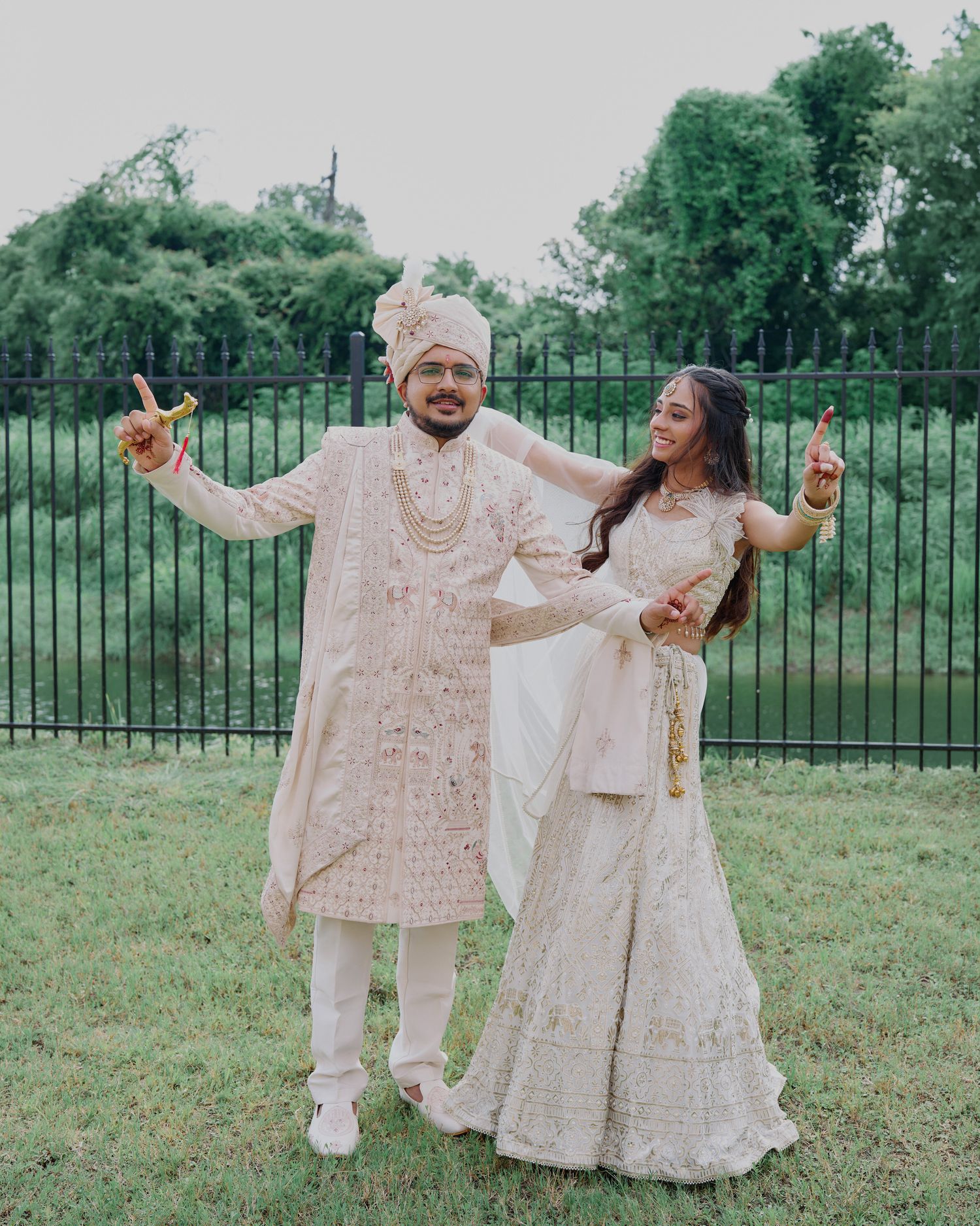 Indian couple in matching cream wedding attire posing joyfully outdoors near a black fence.