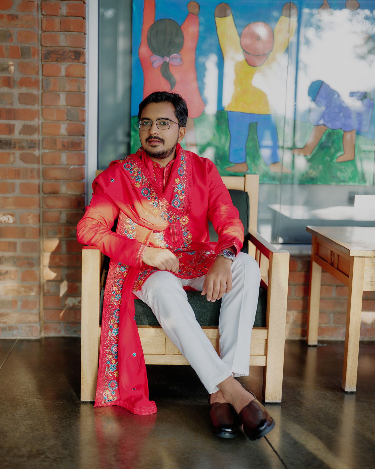 Indian groom in red embroidered sherwani seated in wooden chair before colorful mural wall portrait.