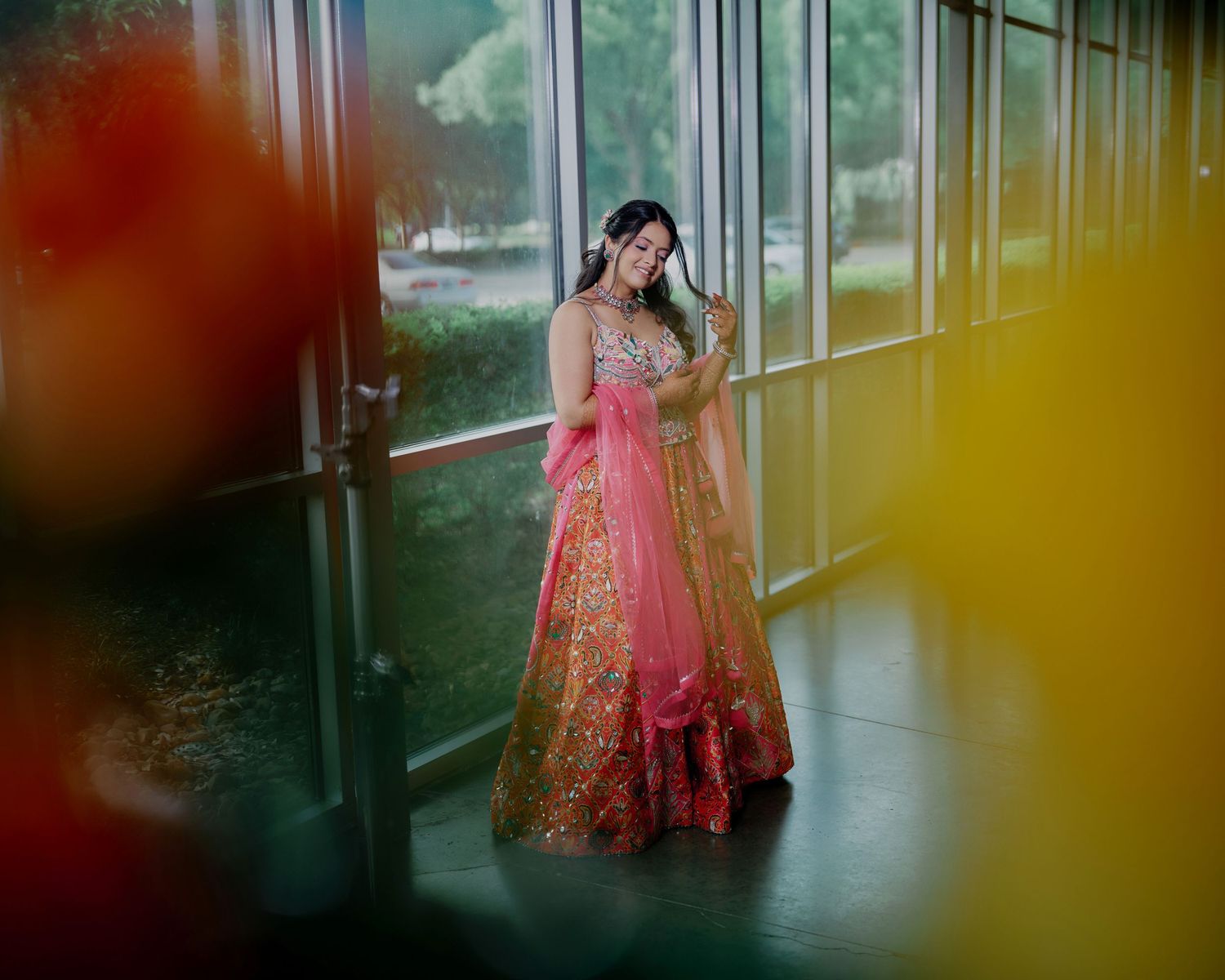 Indian bride in pink and gold lehenga poses gracefully in a sunlit corridor with colorful bokeh.