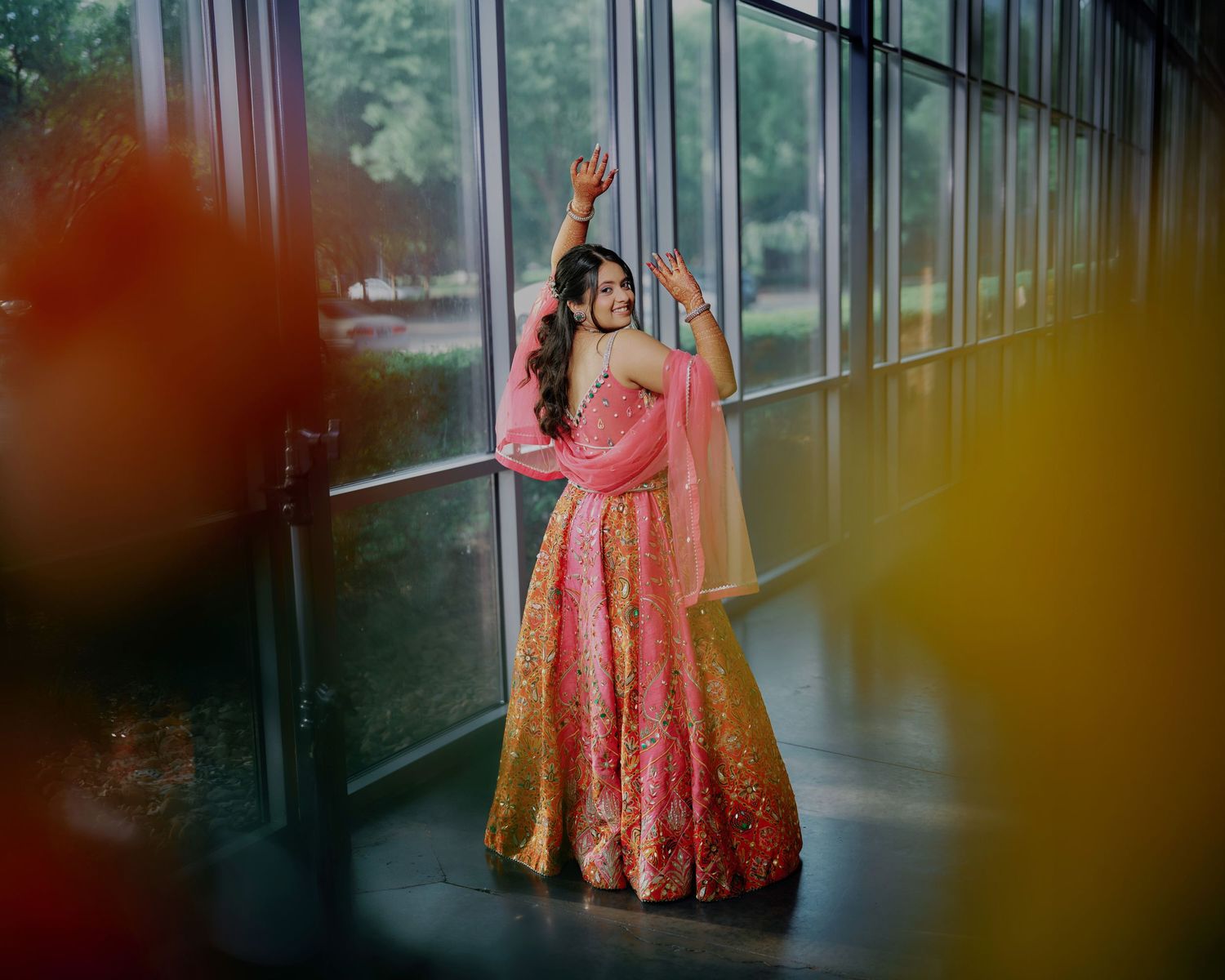 Indian bride in pink and gold lehenga dancing joyfully by large windows in bridal portrait.