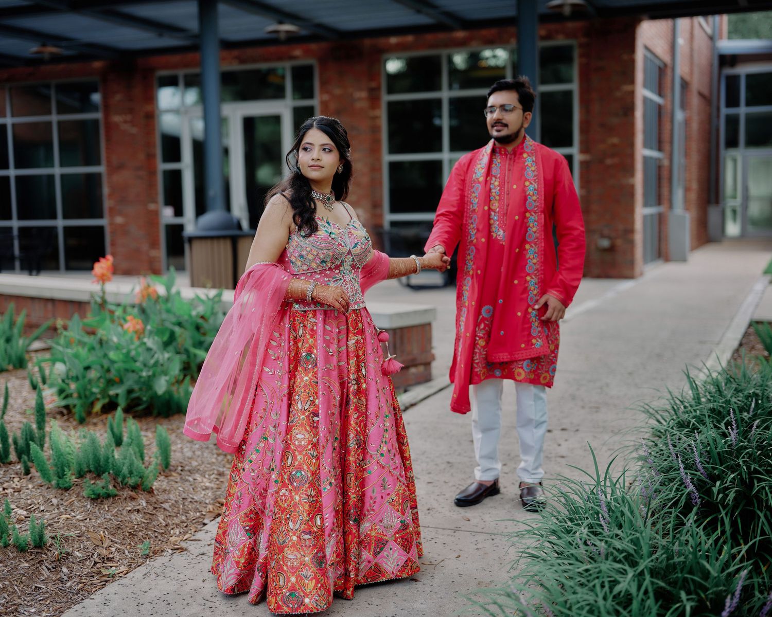 Couple in coordinating pink Indian wedding outfits walking through an outdoor garden setting.