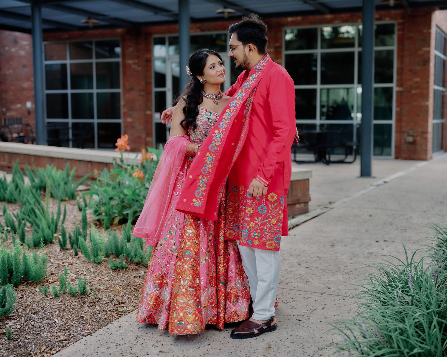 Indian couple in matching pink outfits sharing romantic moment in garden during wedding celebration.