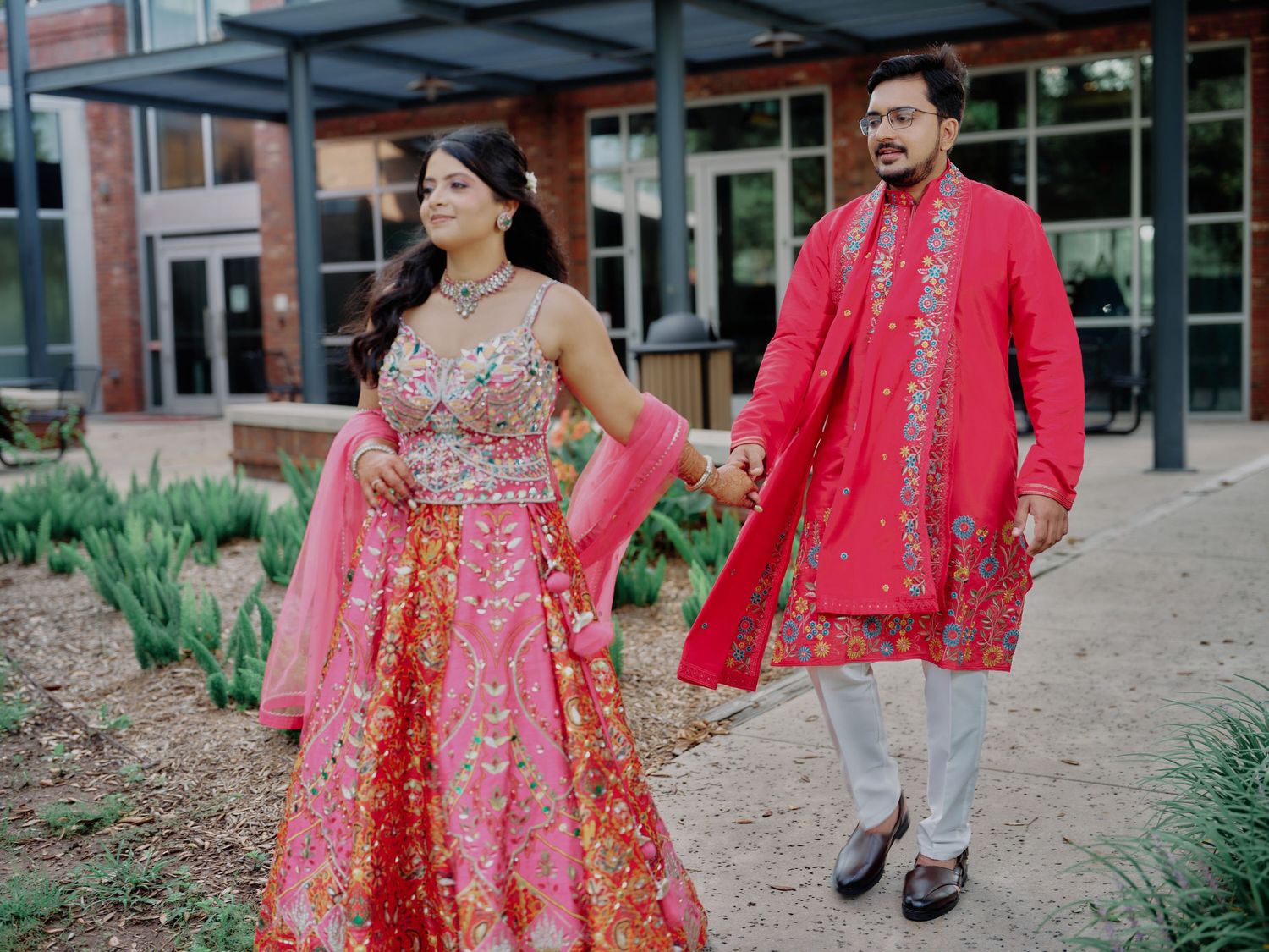 Indian couple in coordinating pink outfits walk hand-in-hand at outdoor wedding venue celebration.