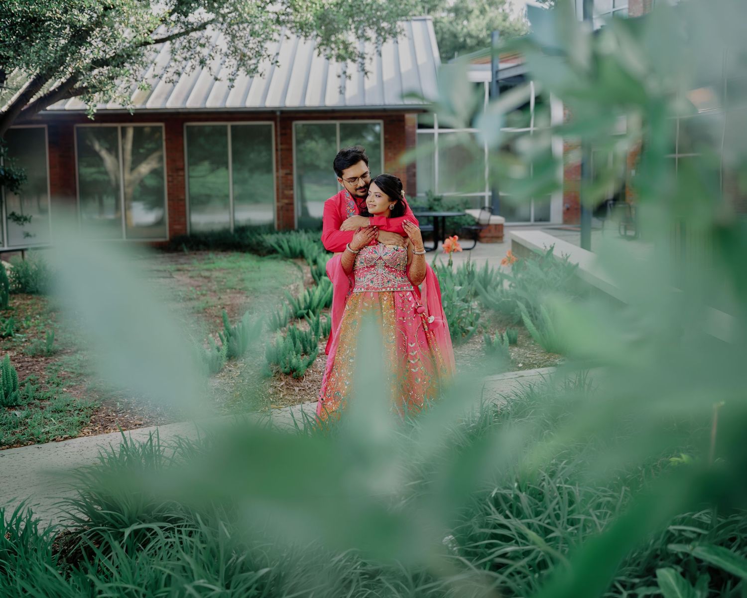 Indian couple in red traditional attire embracing in a lush garden setting.
