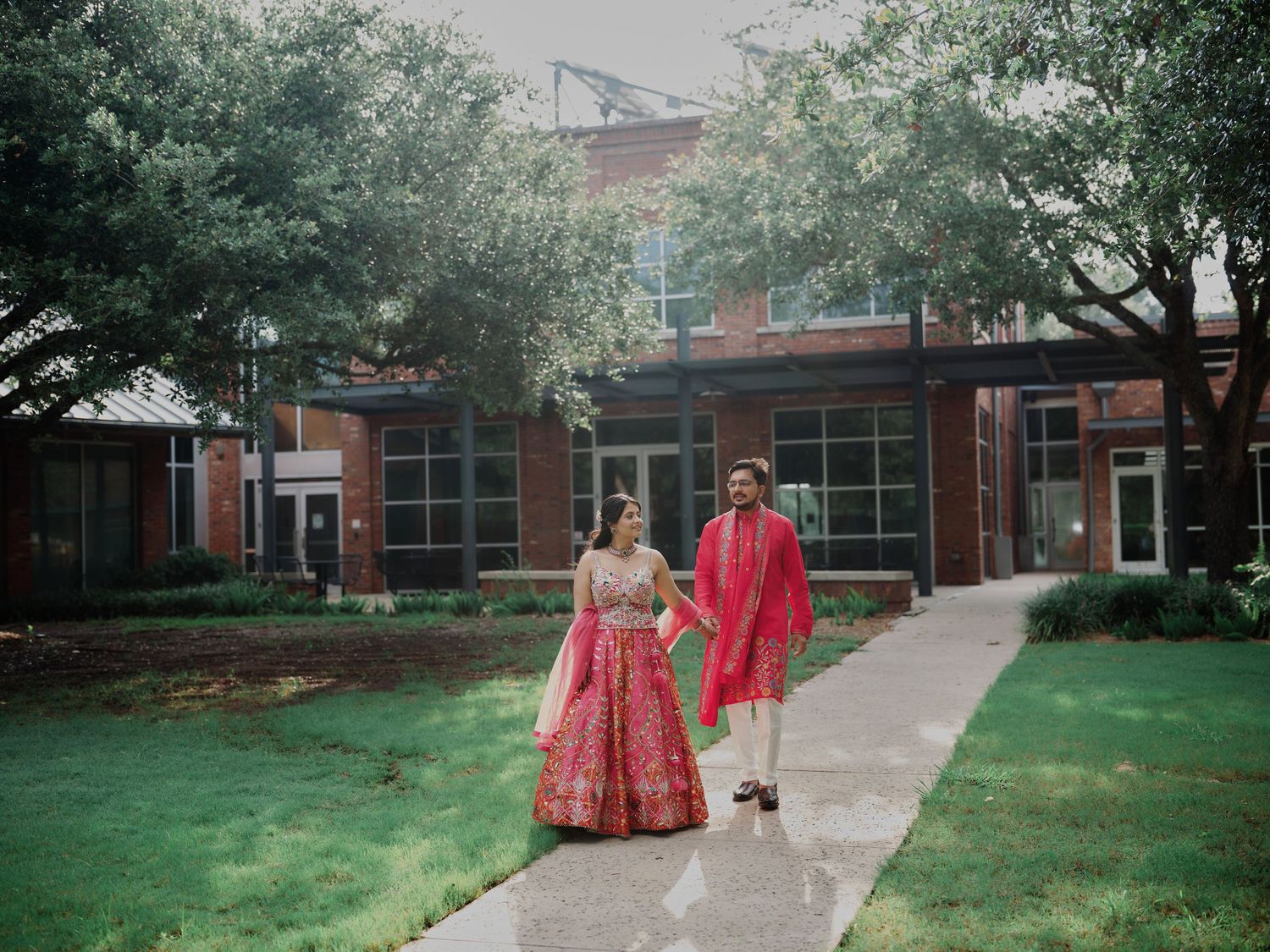 Indian couple in red traditional attire walking on a garden path with lush trees and a brick building backdrop.