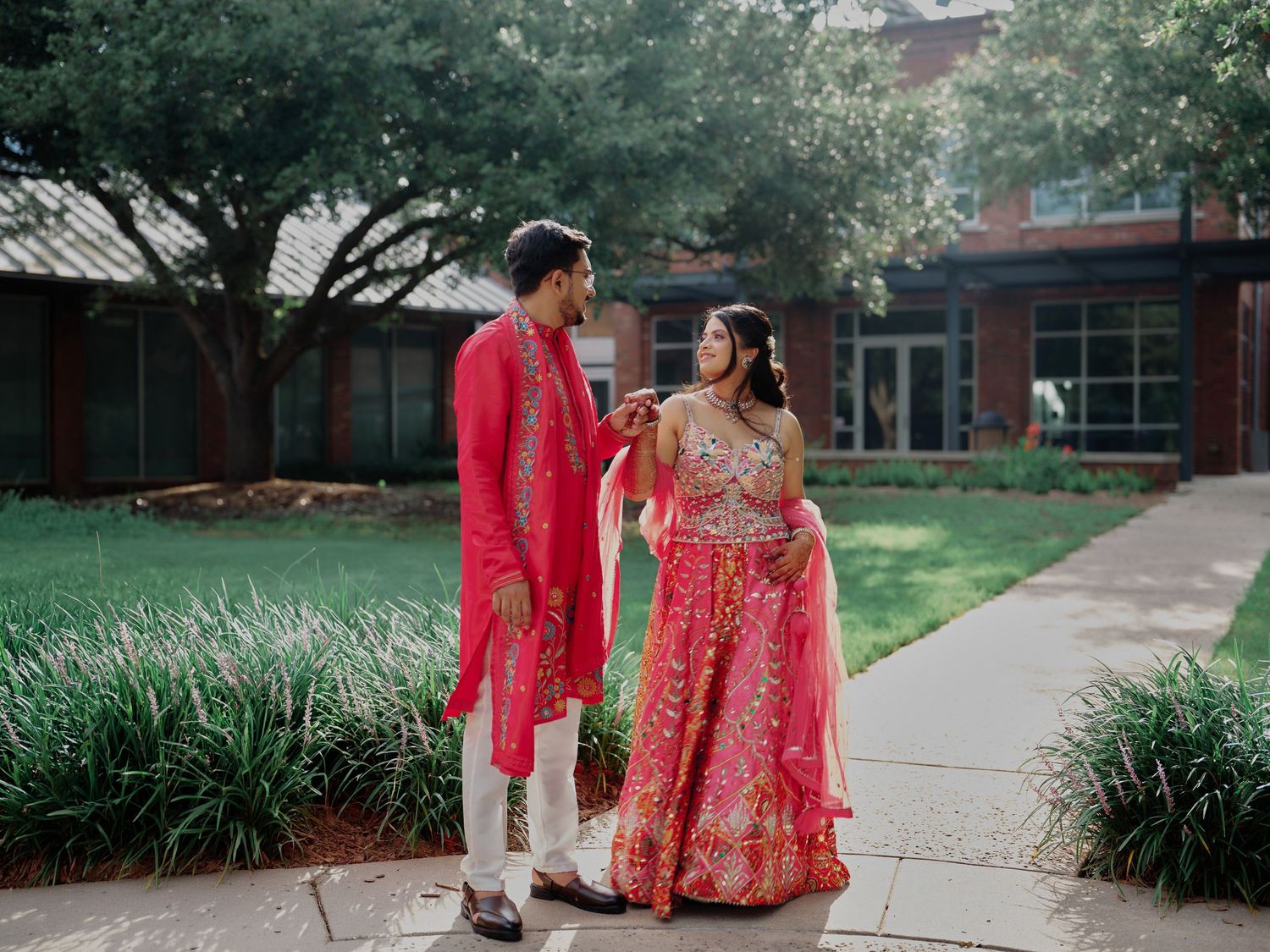 Couple in matching red Indian wedding attire walking outdoors near a brick building with lush greenery.
