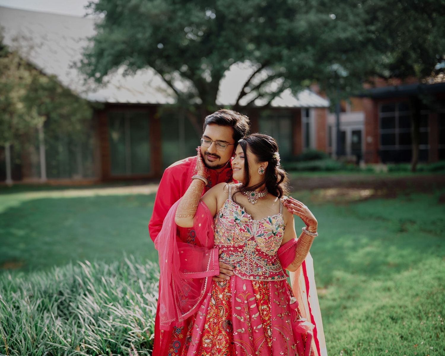 Couple in matching red Indian wedding attire embracing outdoors on green lawn with trees and building backdrop.