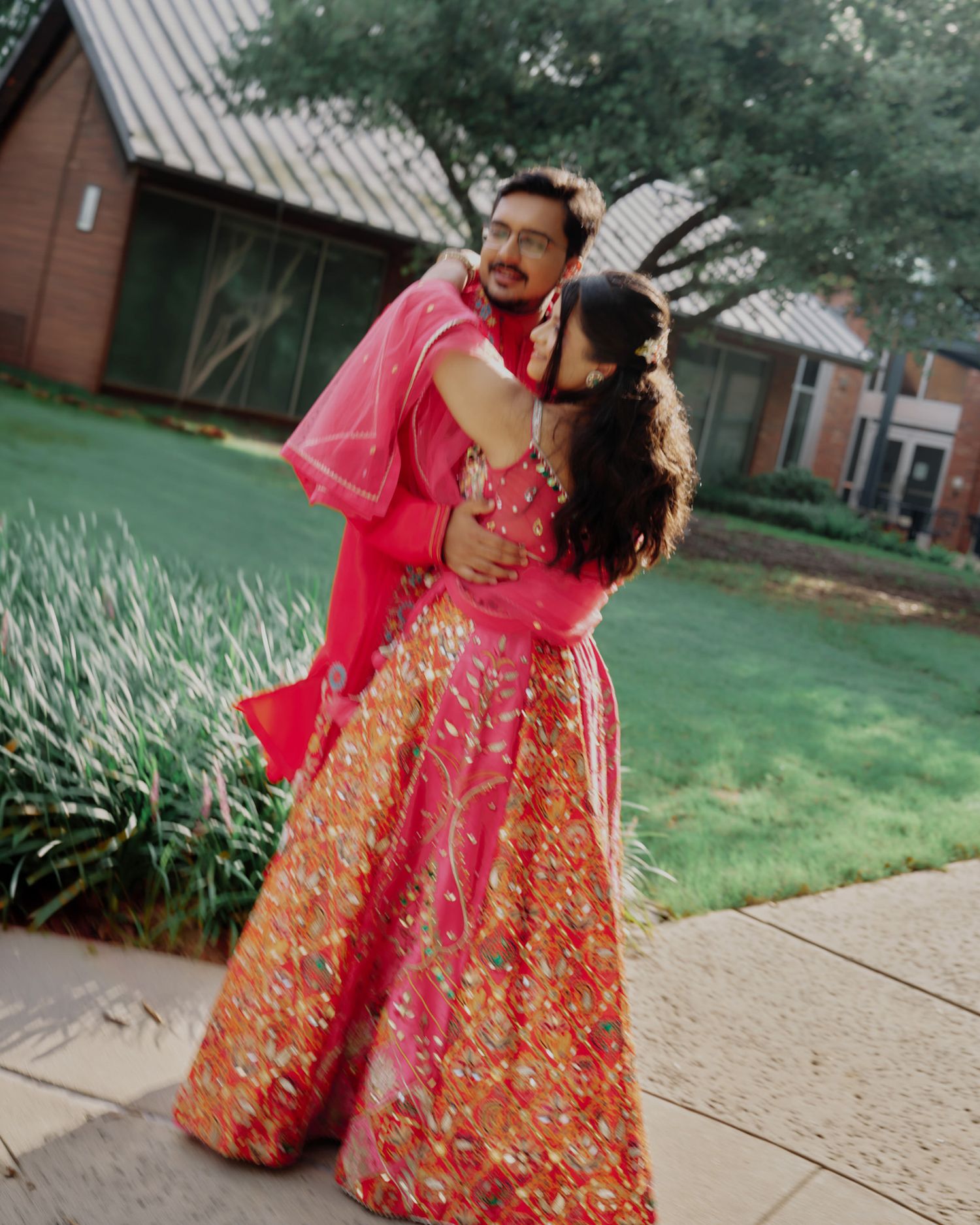 Couple in vibrant pink and orange floral lehenga sharing romantic embrace on outdoor pathway at wedding venue.