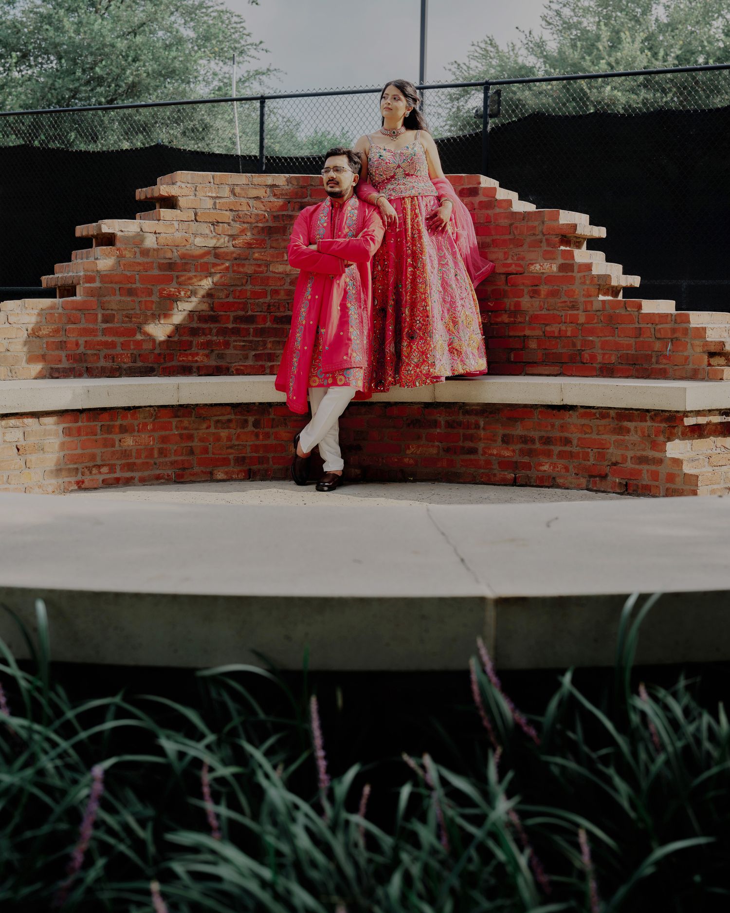 Couple in vibrant pink Indian wedding attire posing on brick steps outdoors.