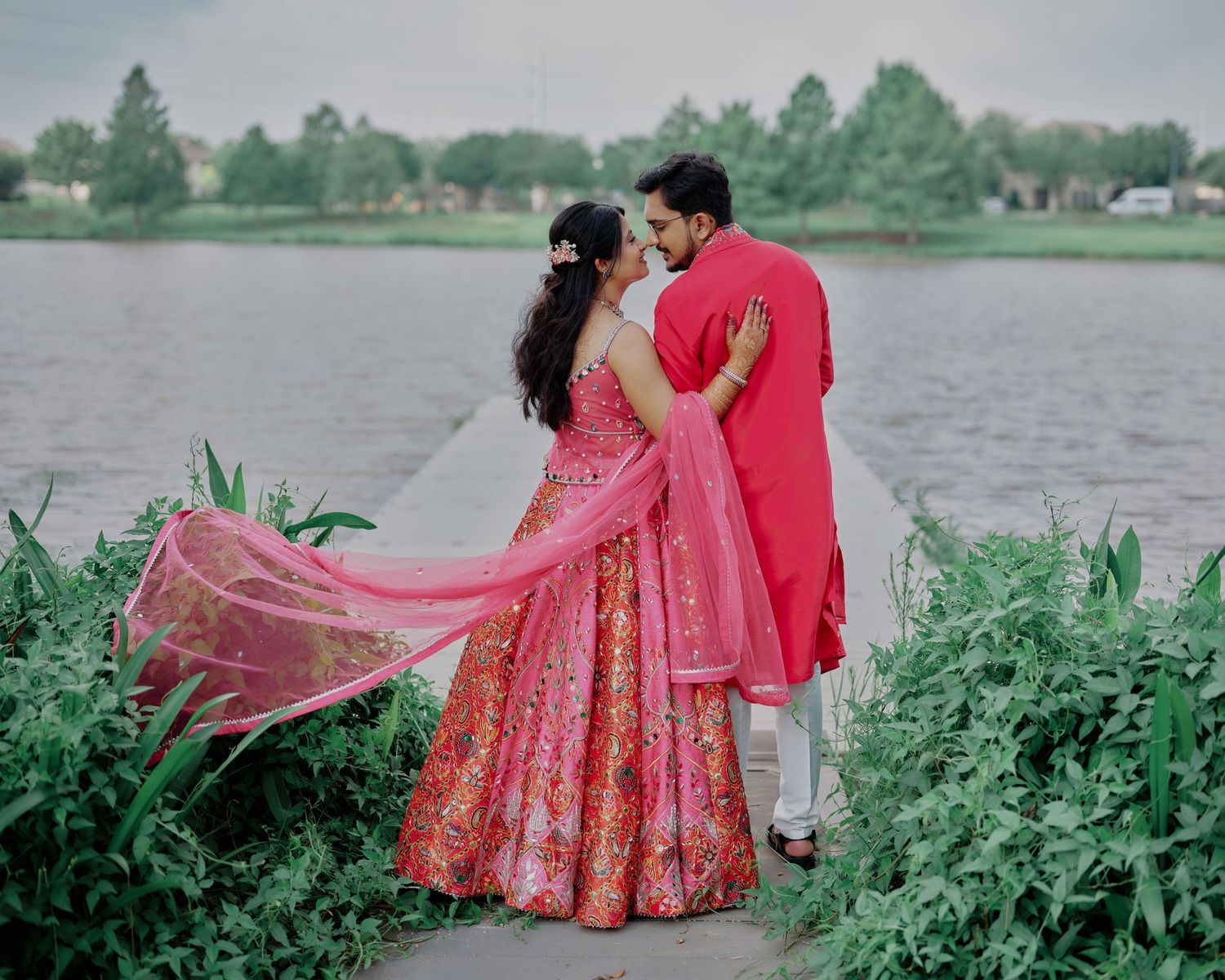 Couple in vibrant red Indian wedding attire sharing a romantic moment by a serene lakeside backdrop.