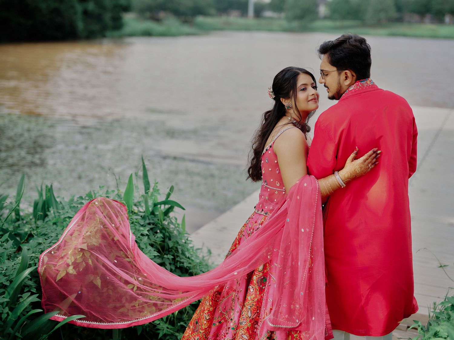 Couple in matching pink Indian attire embracing near a lake, woman's lehenga flowing in the breeze.