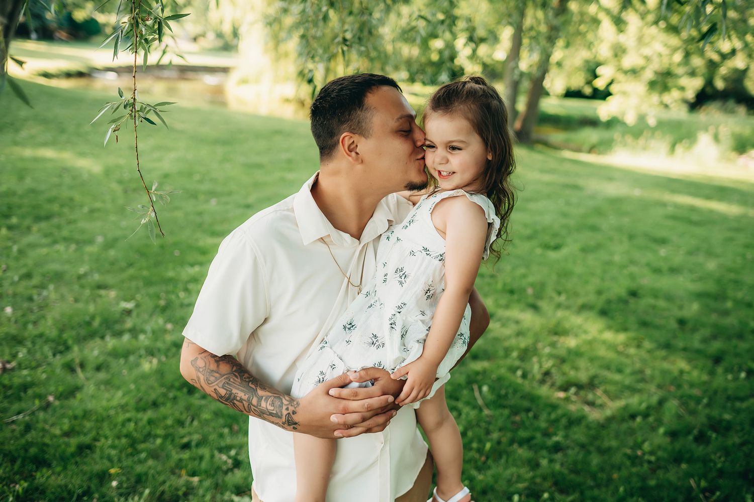 A father lovingly kisses his smiling daughter while holding her in a lush green park on a sunny day.