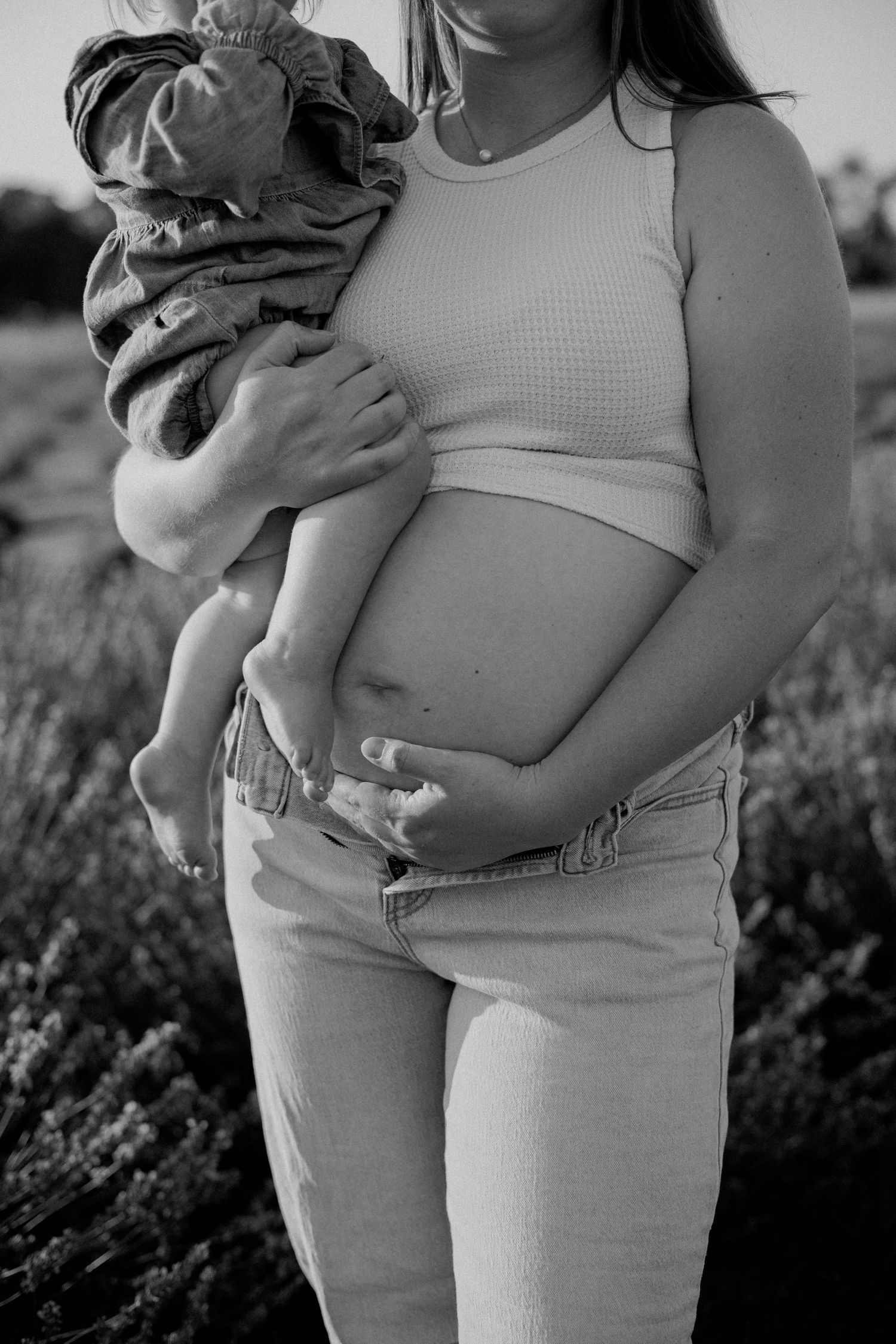 Black and white portrait of pregnant woman holding toddler while cradling baby bump in lavender field.