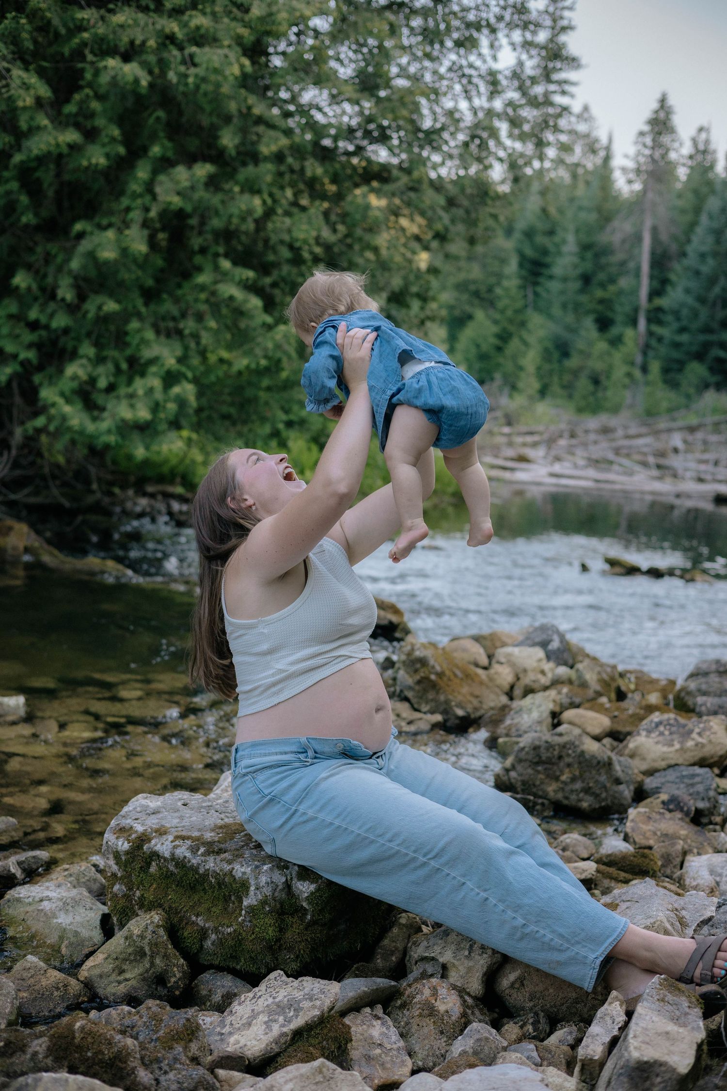 Pregnant woman joyfully lifting smiling baby while sitting on rocks beside a forest river.