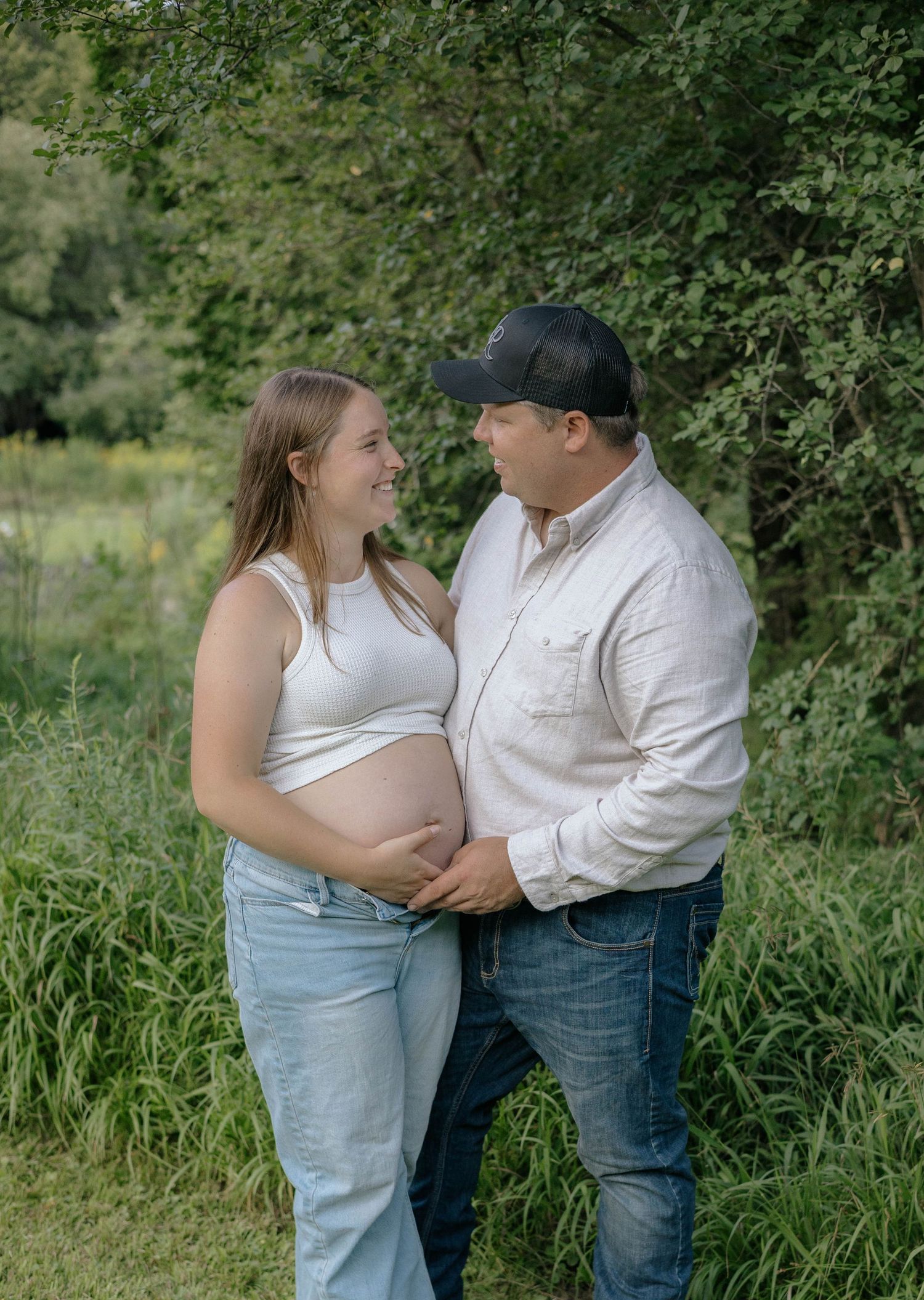 Couple smiling at each other outdoors, woman showing pregnant belly in white crop top and jeans.