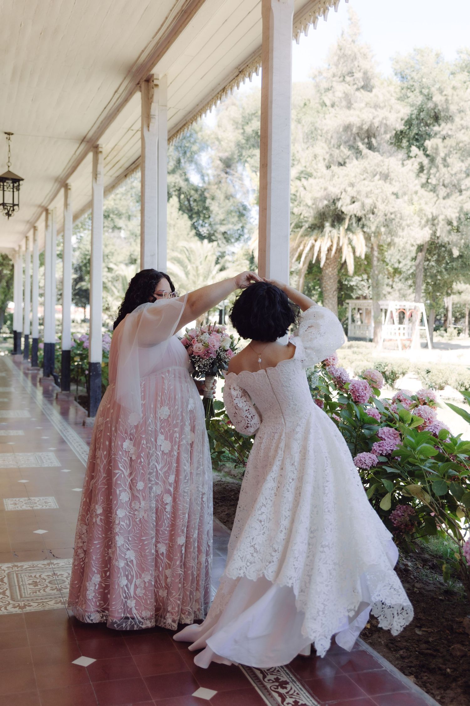 Two brides in lace wedding gowns adjusting hair on elegant garden venue colonnade.