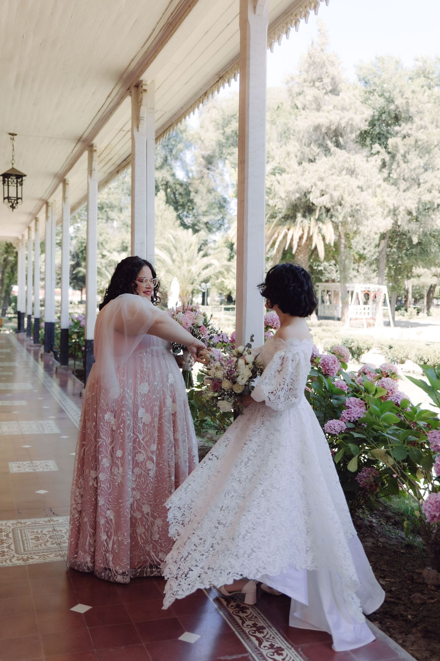 Two brides in white and blush lace gowns exchanging bouquet on beautiful garden colonnade.