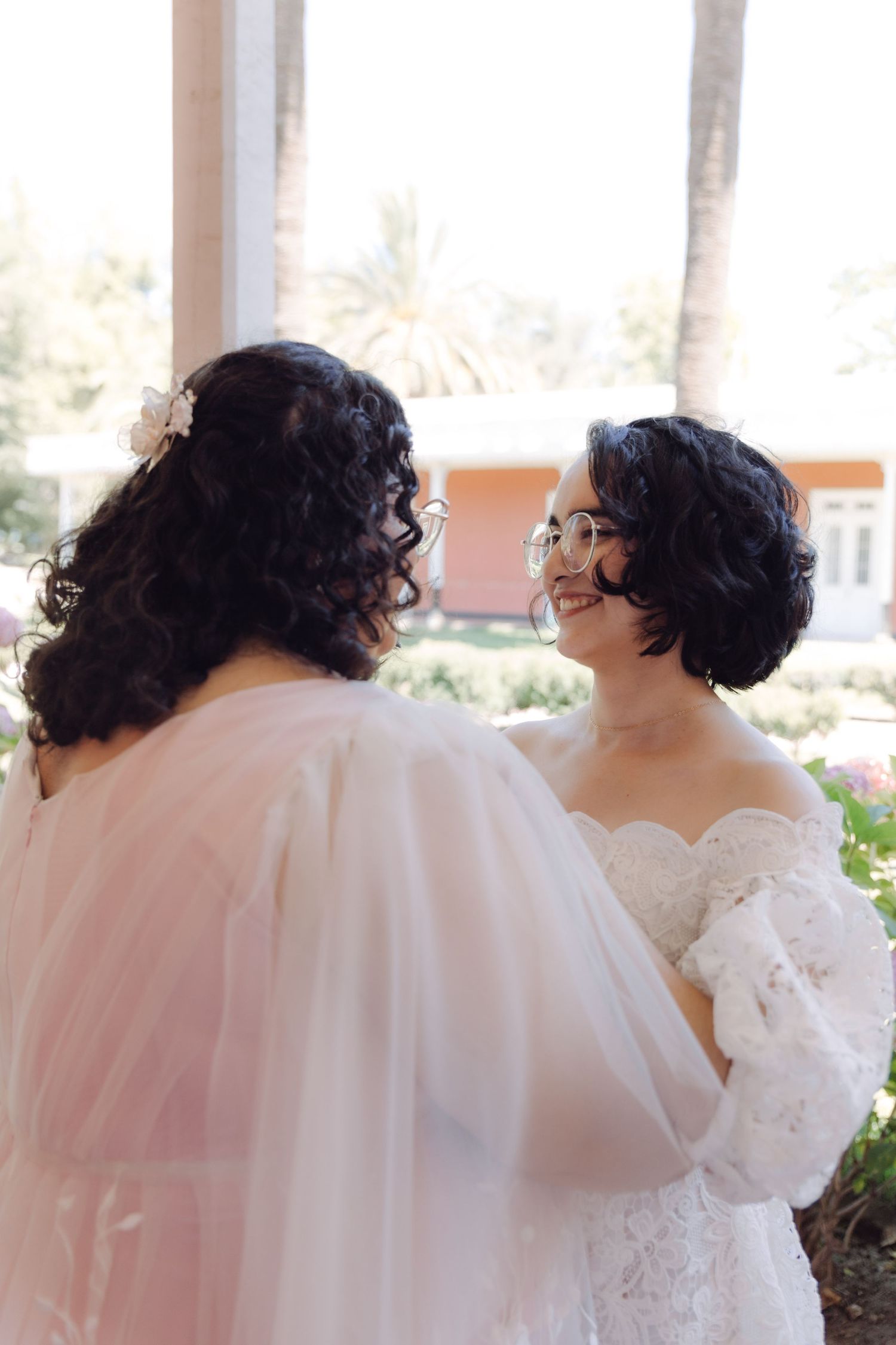 Two brides in white and blush lace wedding gowns sharing joyful moment at garden venue.