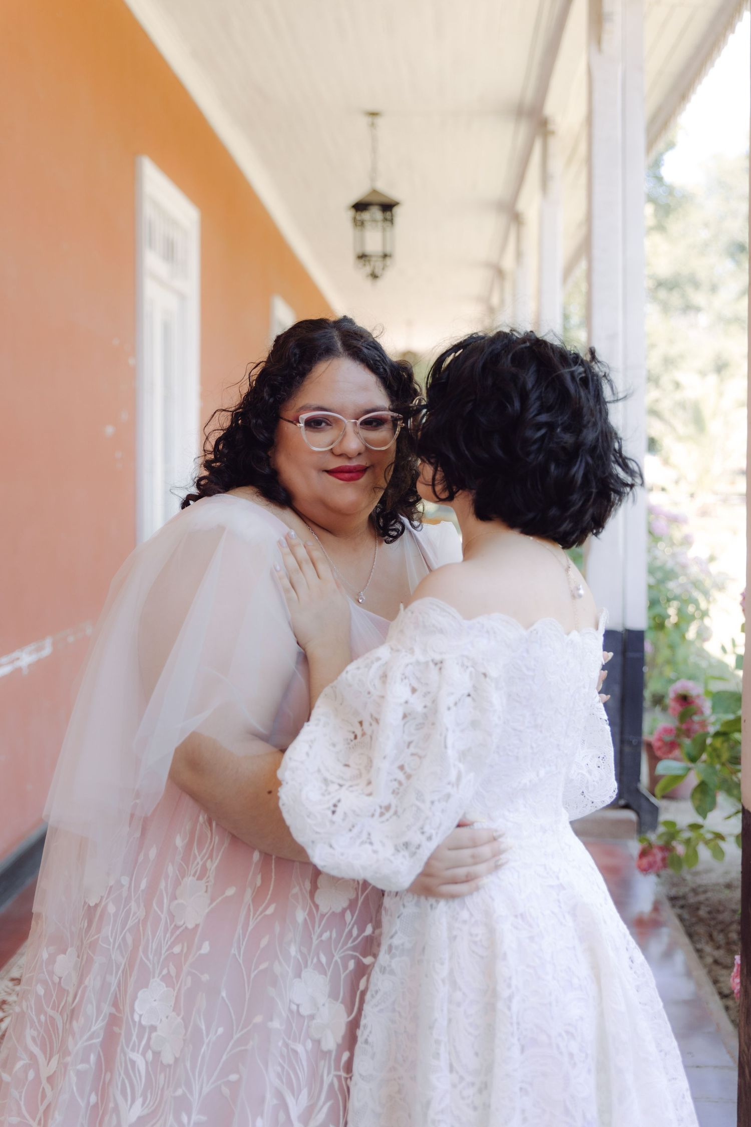 Two brides in white wedding dresses embrace on a pink building porch, one wearing glasses and a veil.