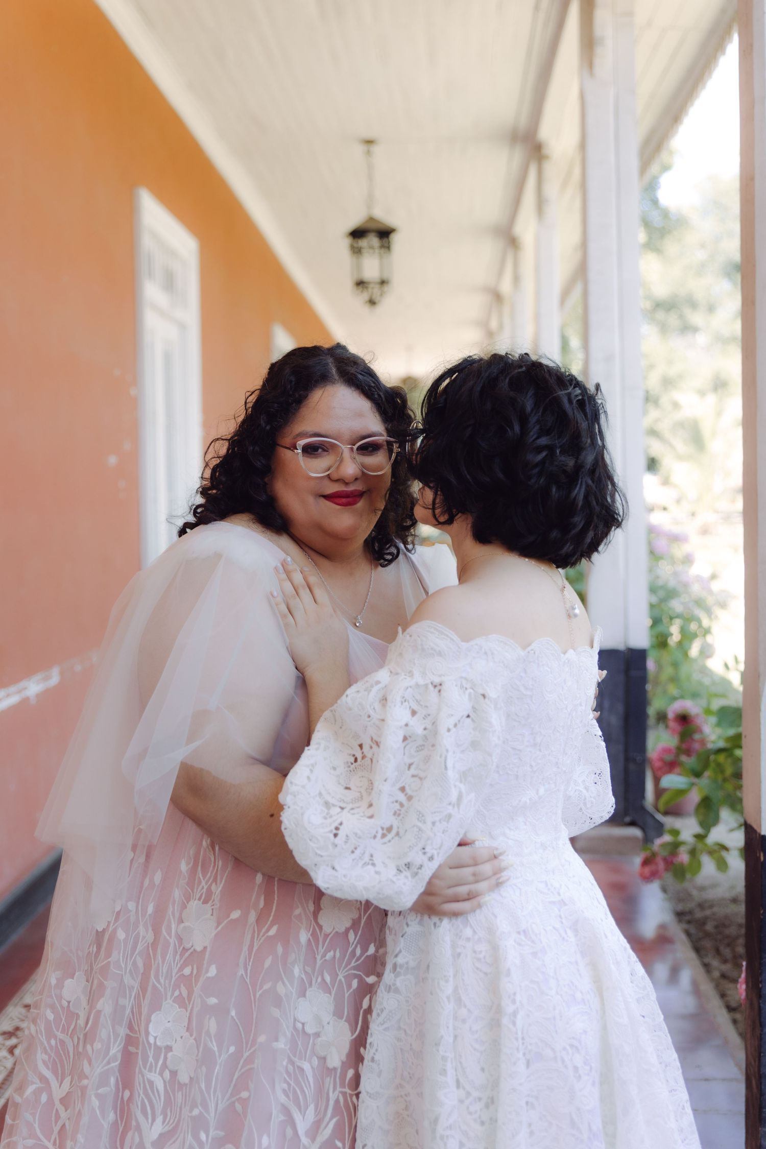 Two brides in lace wedding gowns share an intimate embrace on a sunlit colonial-style porch.