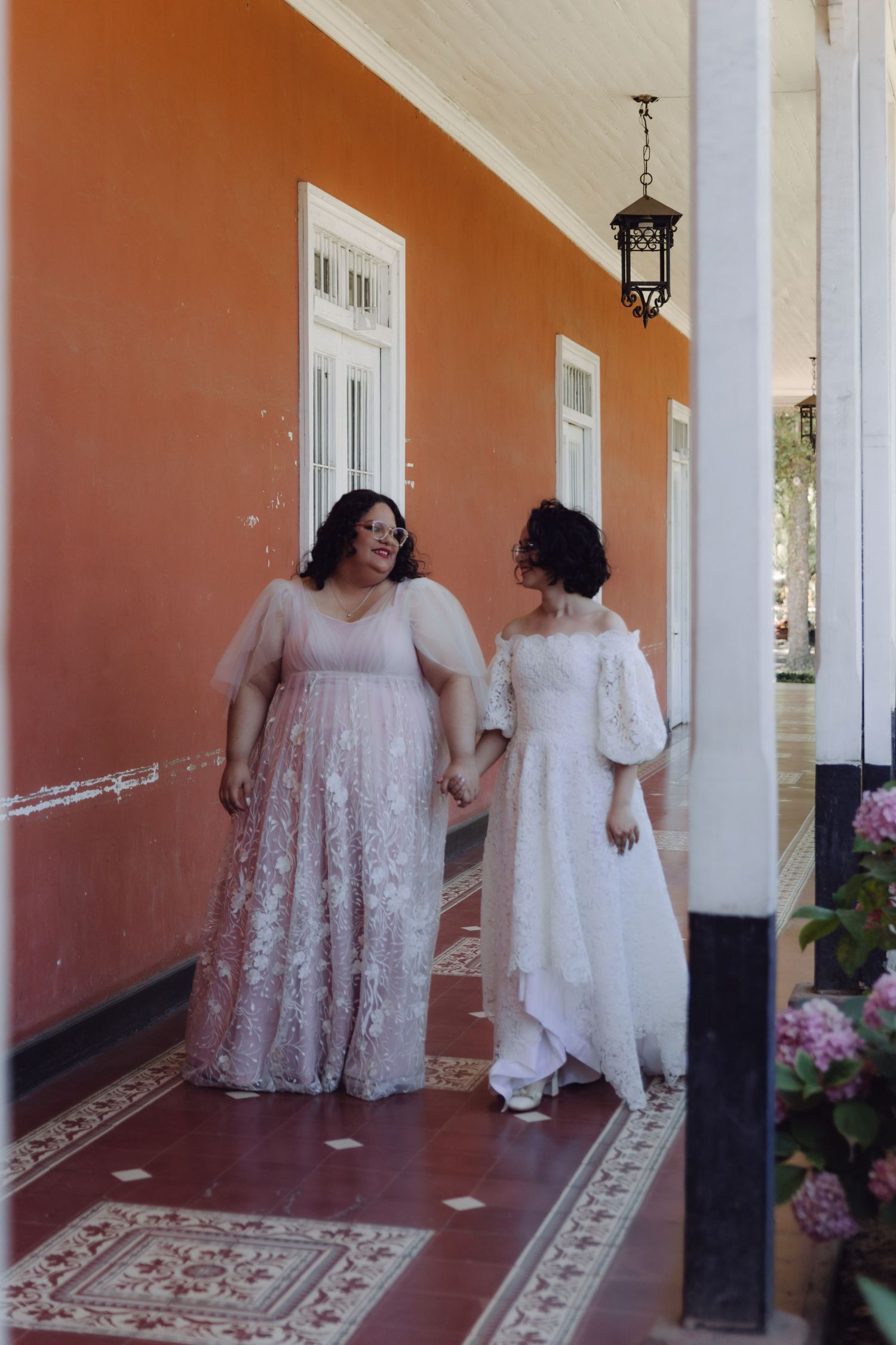 Two brides in elegant white and blush wedding dresses walk hand in hand along a terracotta porch.