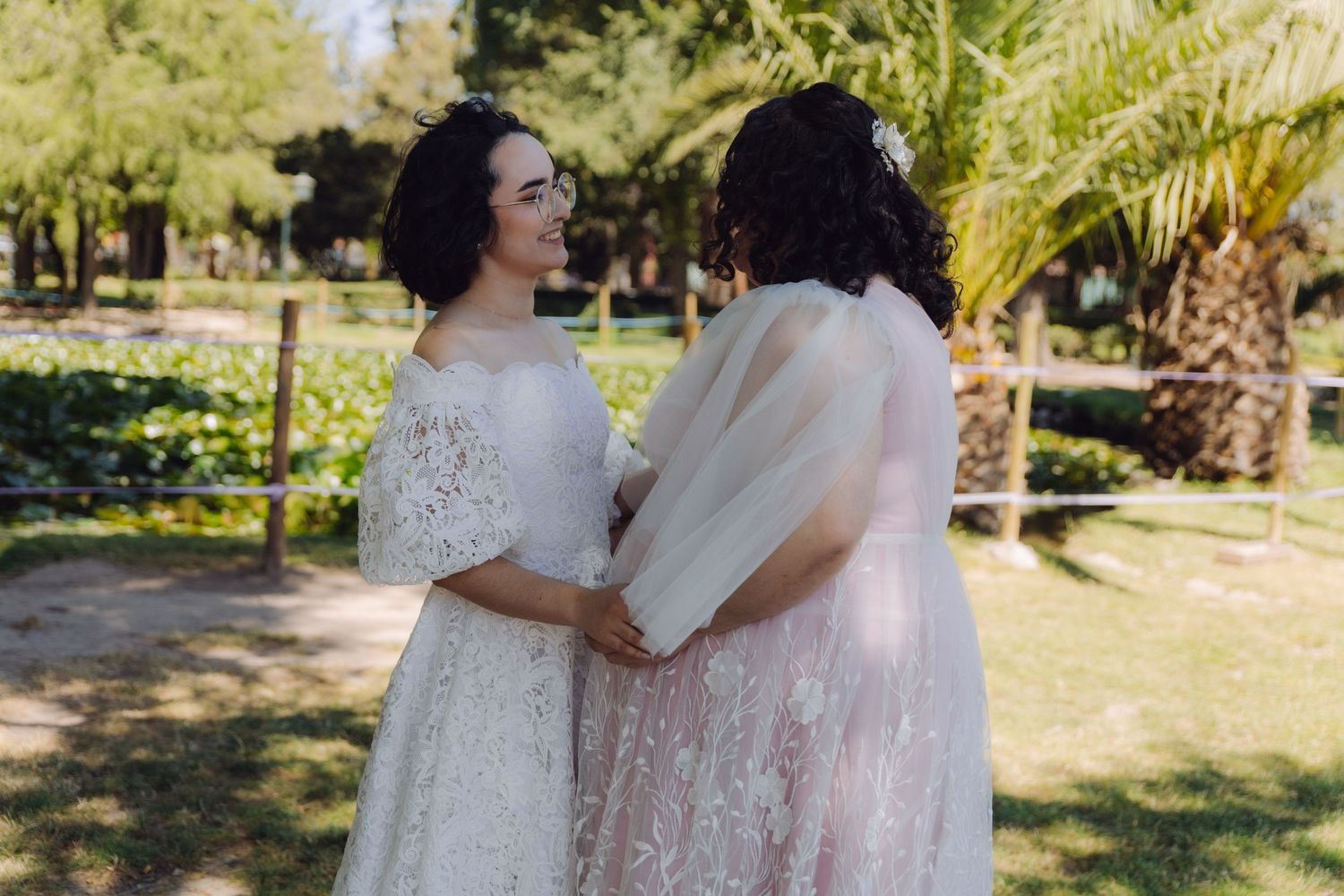 Two brides in white lace wedding dresses hold hands and smile in a sunny garden setting.