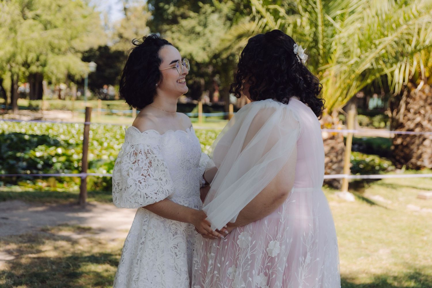 Two brides in white lace wedding gowns face each other holding hands in a lush garden.