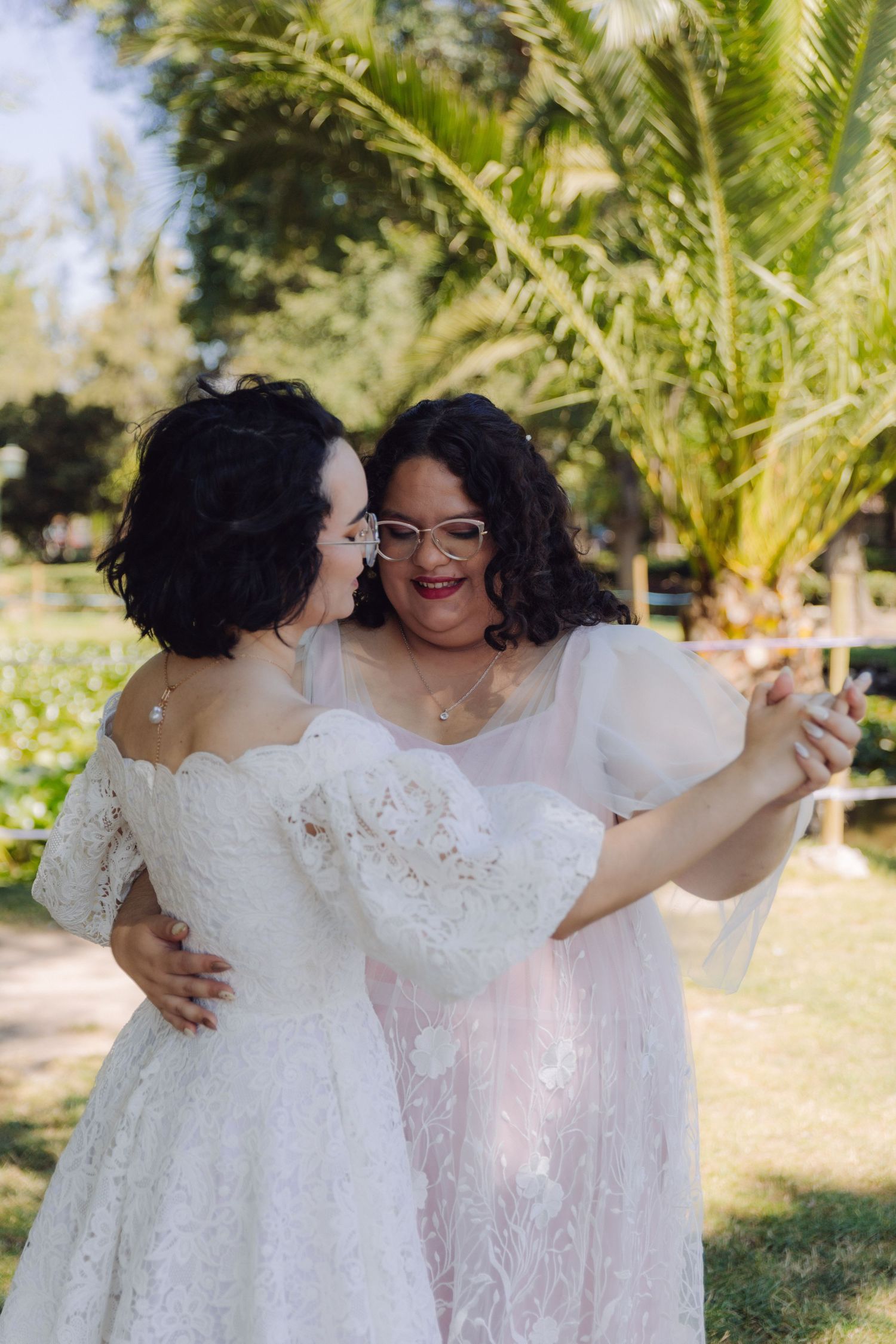 Two brides in white lace wedding dresses embrace and laugh together in a sunny garden.